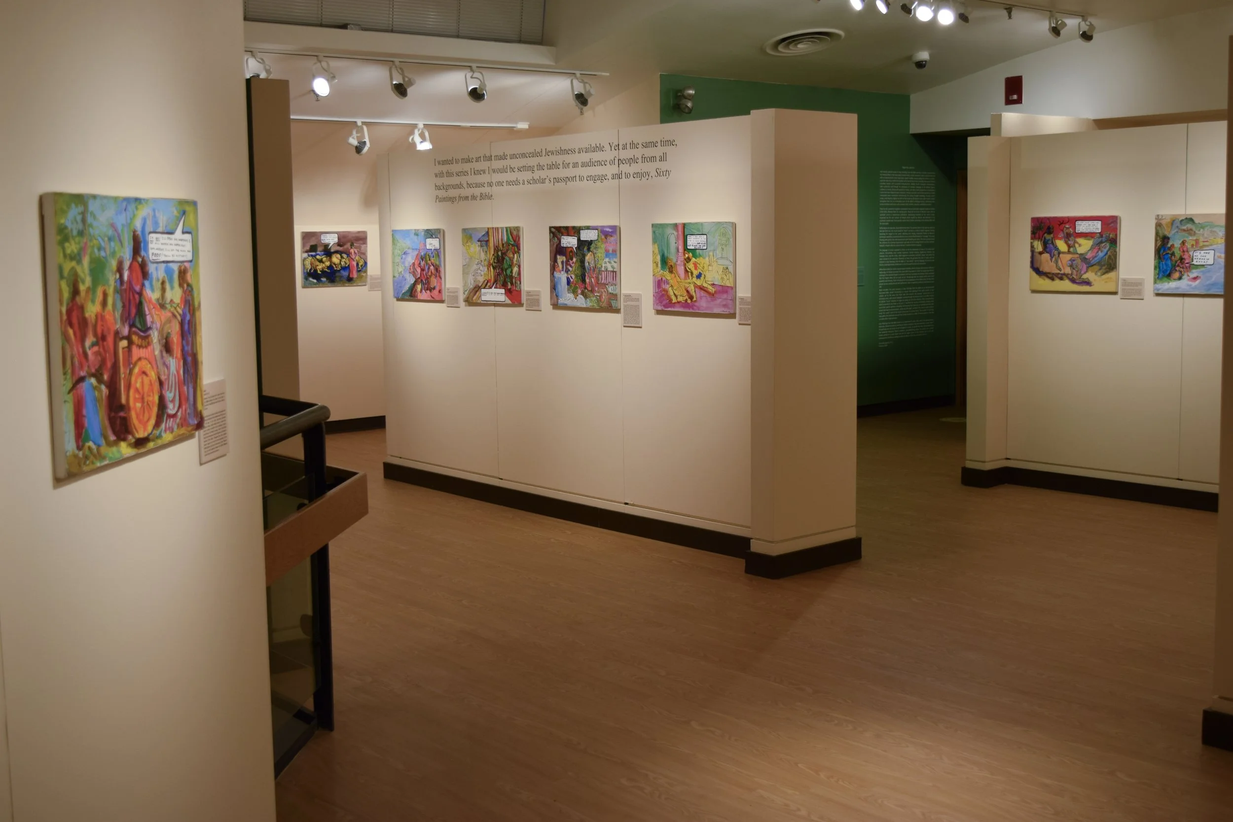 An art gallery featuring colorful paintings displayed on white walls with spotlights overhead. The paintings depict various scenes and are accompanied by descriptive plaques.