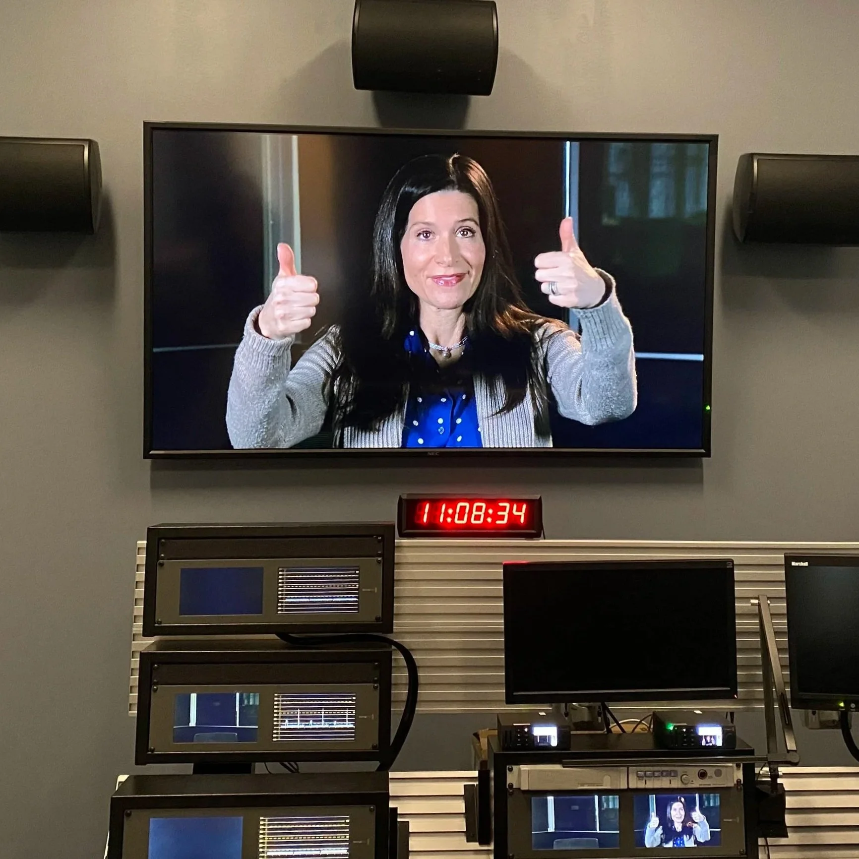 Samantha Baskind on a television screen giving two thumbs up, in a control room with monitors and a digital clock reading 11:08:34.