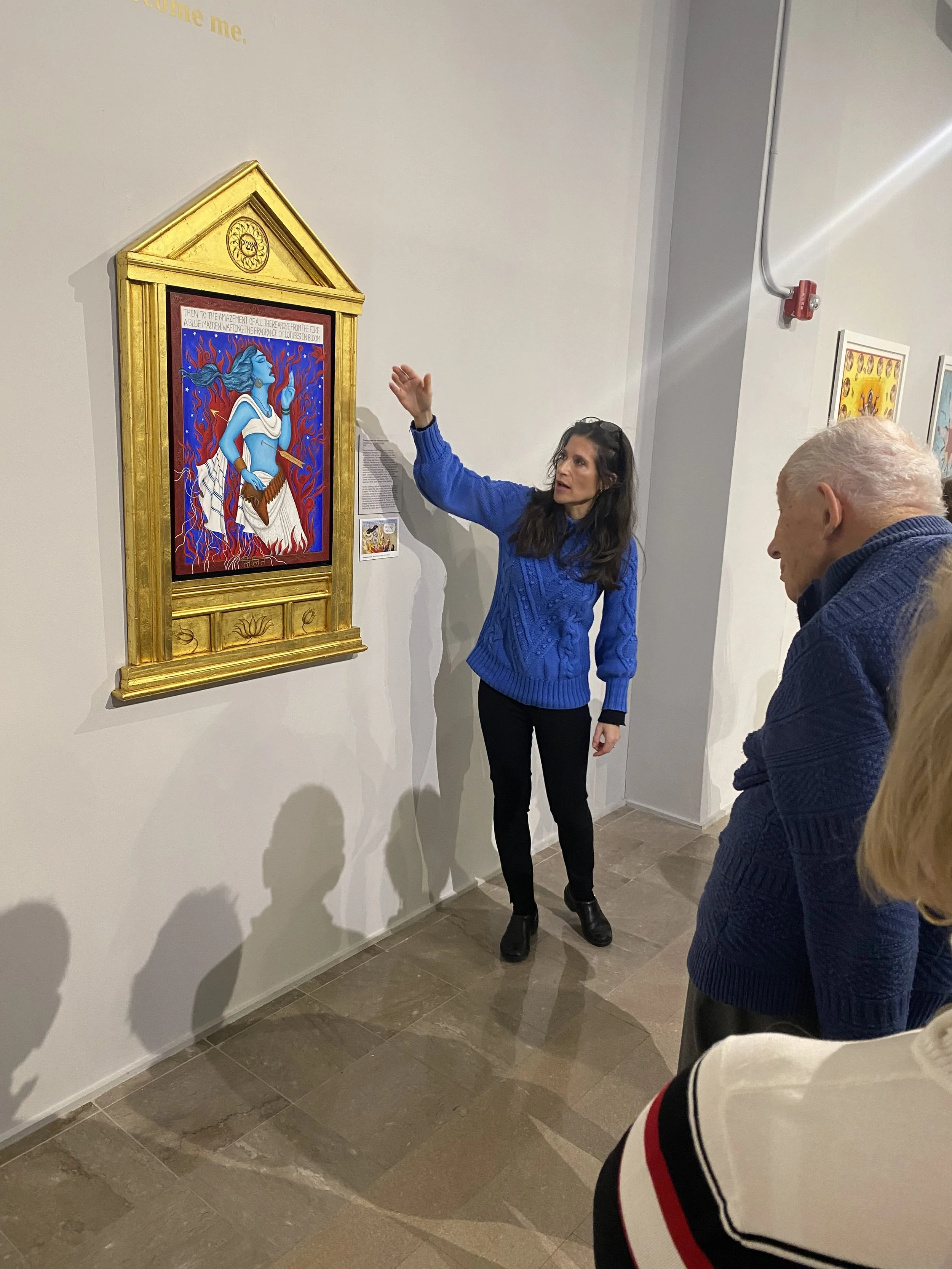 Samantha Baskind explaining a colorful painting of a blue-skinned woman with blue hair, surrounded by red flames, to a group of people in an art gallery.