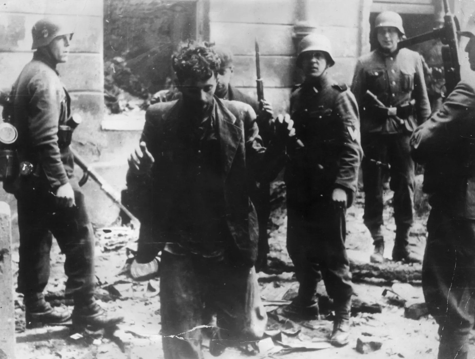 Historical black and white photo of Nazi soldiers and a man who appears to be a prisoner in a destroyed urban area during World War II.