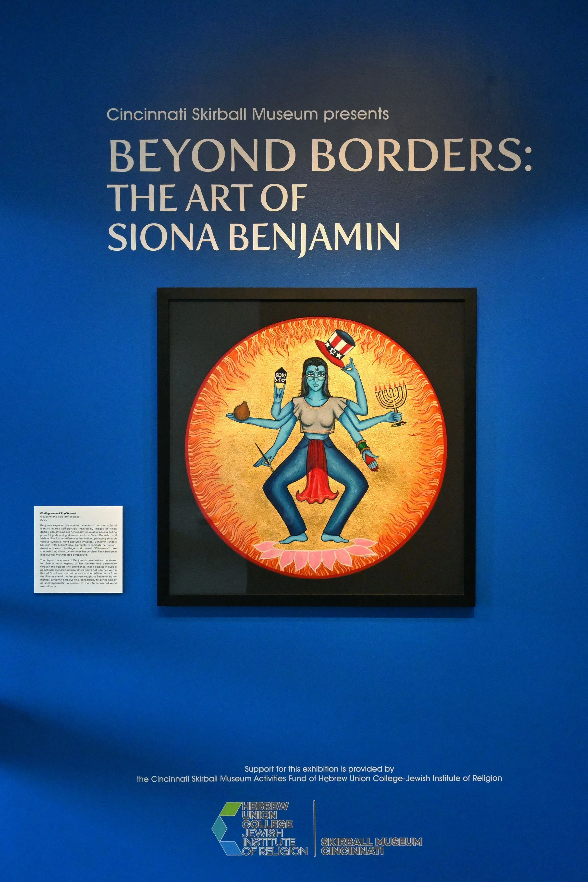 Exhibition poster for 'Beyond Borders: The Art of Siona Benjamin' at the Cincinnati Skirball Museum featuring an artwork of a multi-armed deity with humor-themed accessories.