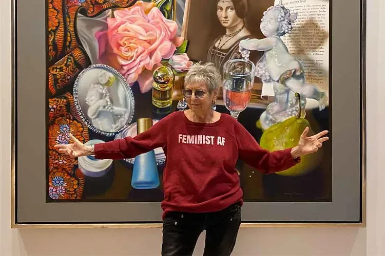 A woman with short gray hair and sunglasses standing in front of a colorful painting. She is wearing a red sweatshirt that says 'Feminist AF' and has her arms spread out.
