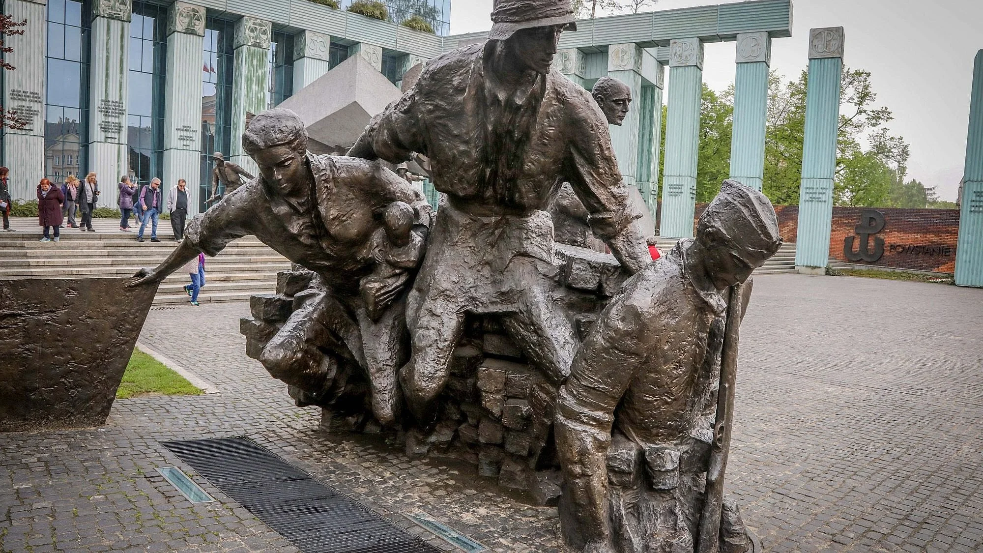 Bronze sculpture of five workers, with some appearing to be breaking rocks or engaged in construction tasks, in front of a modern building with glass and columns, and a group of people walking nearby.