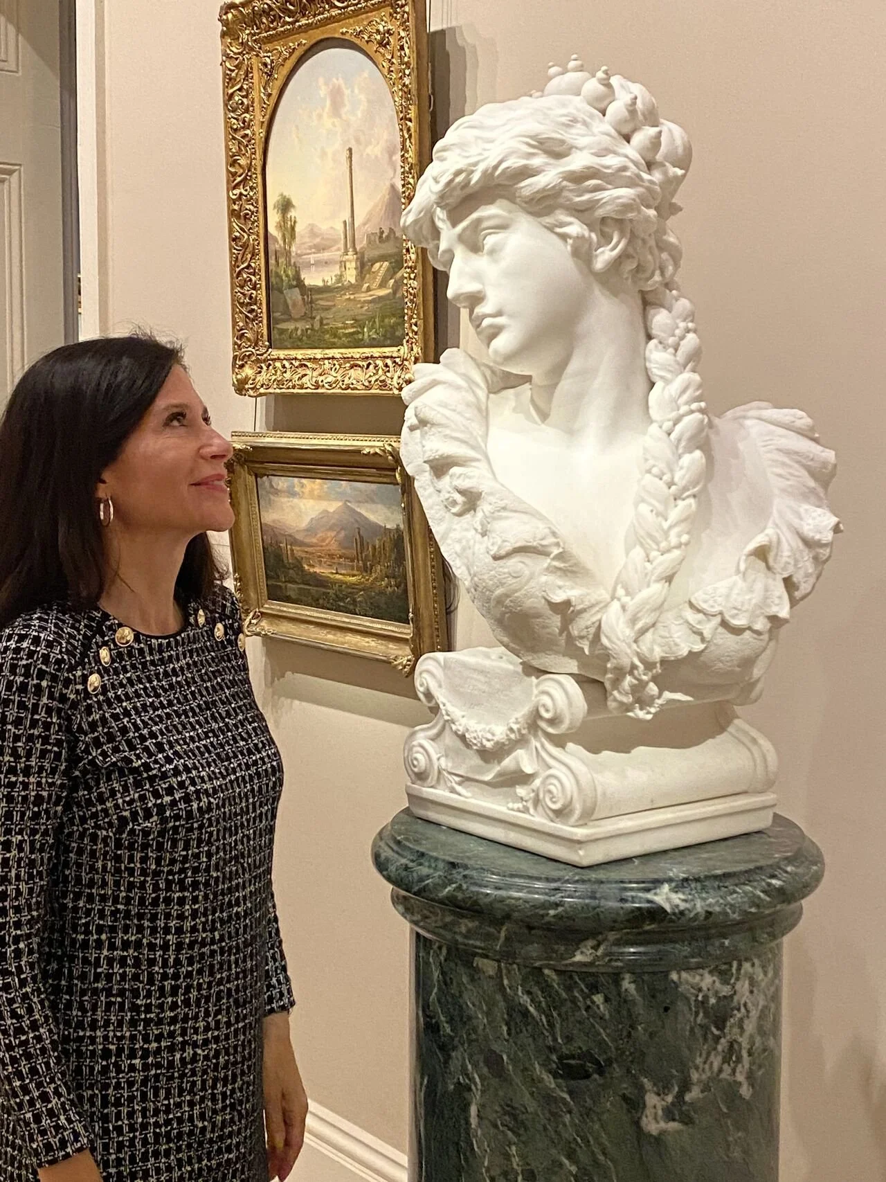 Samantha Baskind looking at a white marble bust of a woman with braided hair, on a green marble pedestal, in an art gallery with landscape paintings in gilded frames behind her.