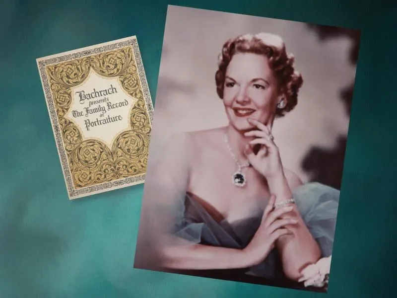 A vintage portrait photograph of a woman with short, curly hair, wearing a strapless dress and jewelry, resting her chin on her hand with a smile. Beside the photograph, there is an ornate card titled "Bachrach presents The Family Record of Portraiture."
