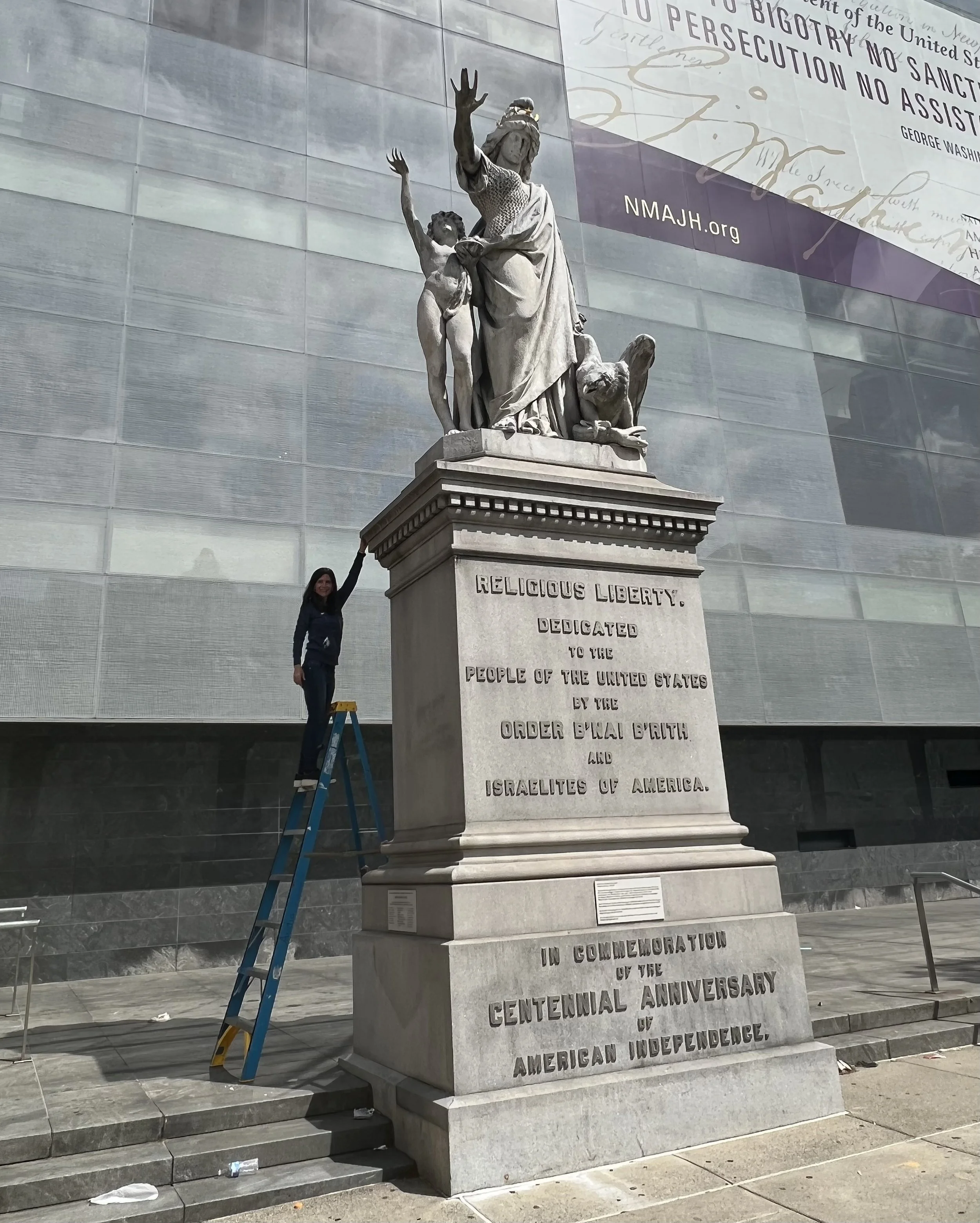 A woman standing on a ladder touching the statue of a female figure on a monument. The monument is dedicated to religious liberty and the dedication to the people of the United States by the order B'nai B'rith and Israelites of America. It commemorates the centennial anniversary of American independence.