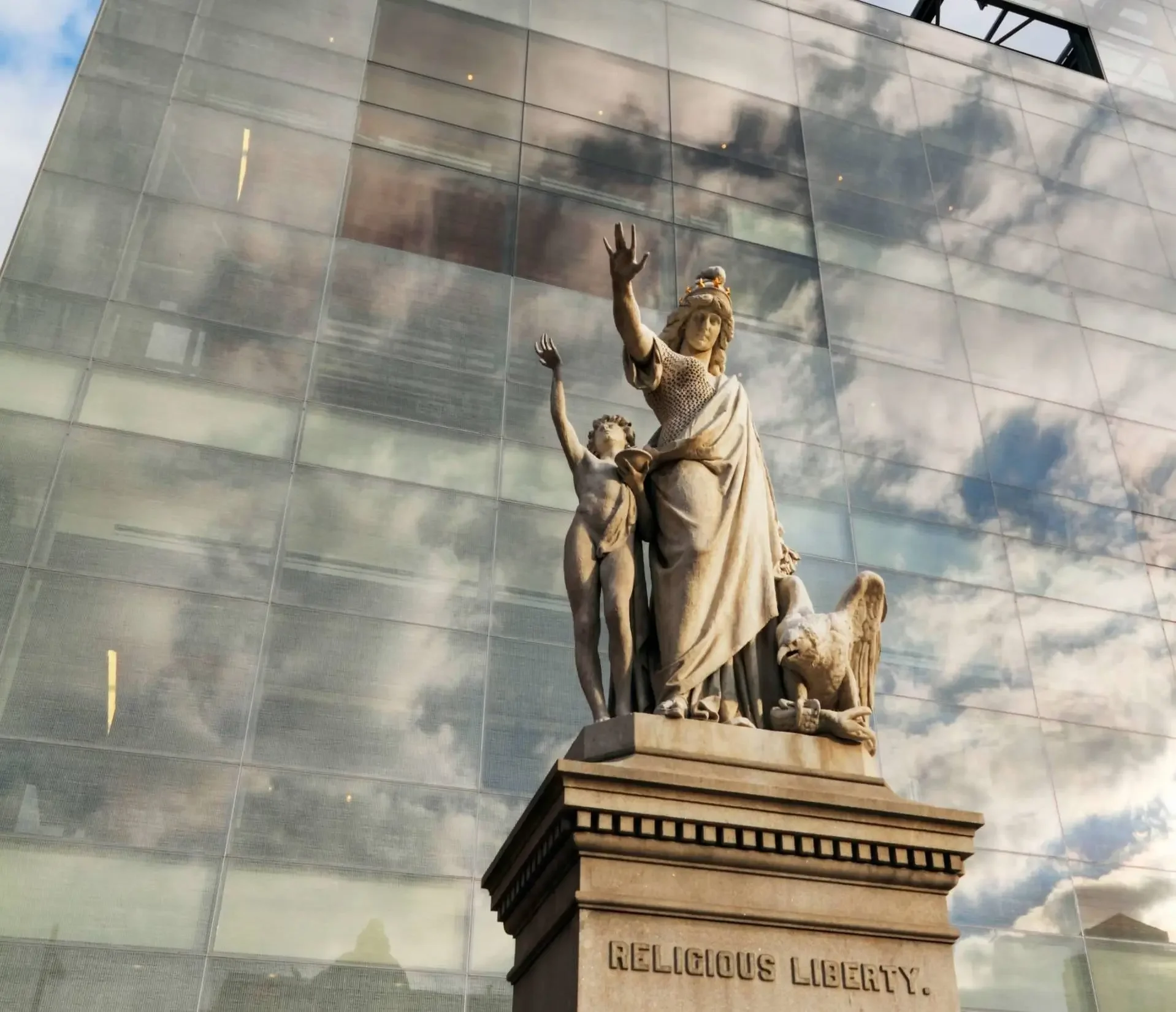 Statue of a woman with a crown and two children, one reaching upward, and a lion, mounted on a pedestal with the inscription "RELIGIOUS LIBERTY", in front of a reflective glass building.