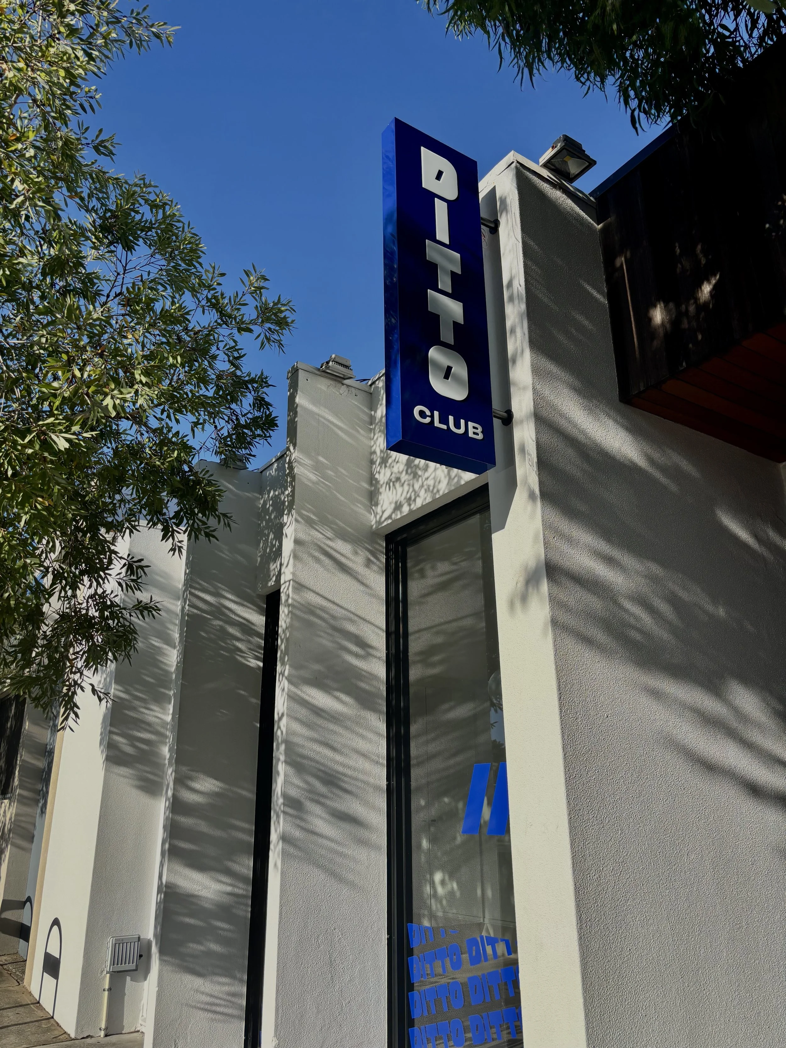 Exterior of a nightclub with a vertical blue sign that reads "PITTO CLUB" attached to a white building with glass windows.