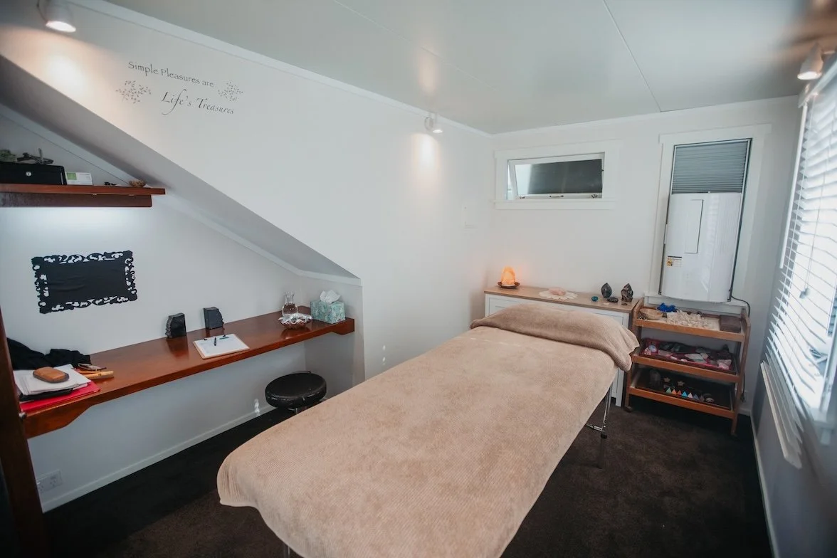Tarla's treatment room with a treatment table covered in a beige sheet, a small white cabinet with Himalayan salt lamp and decorative stones, wooden shelves with crystals, and a small decorative sign on the wall.