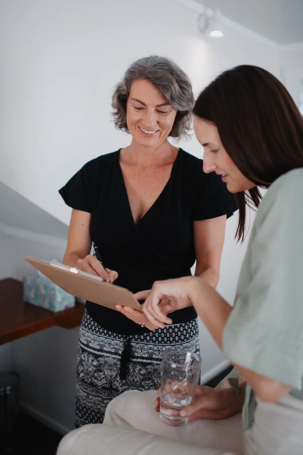 Tarla and her client engaged in a discussion, Tarla holding a clipboard and pen, the client with a glass of water, in a well-lit room.
