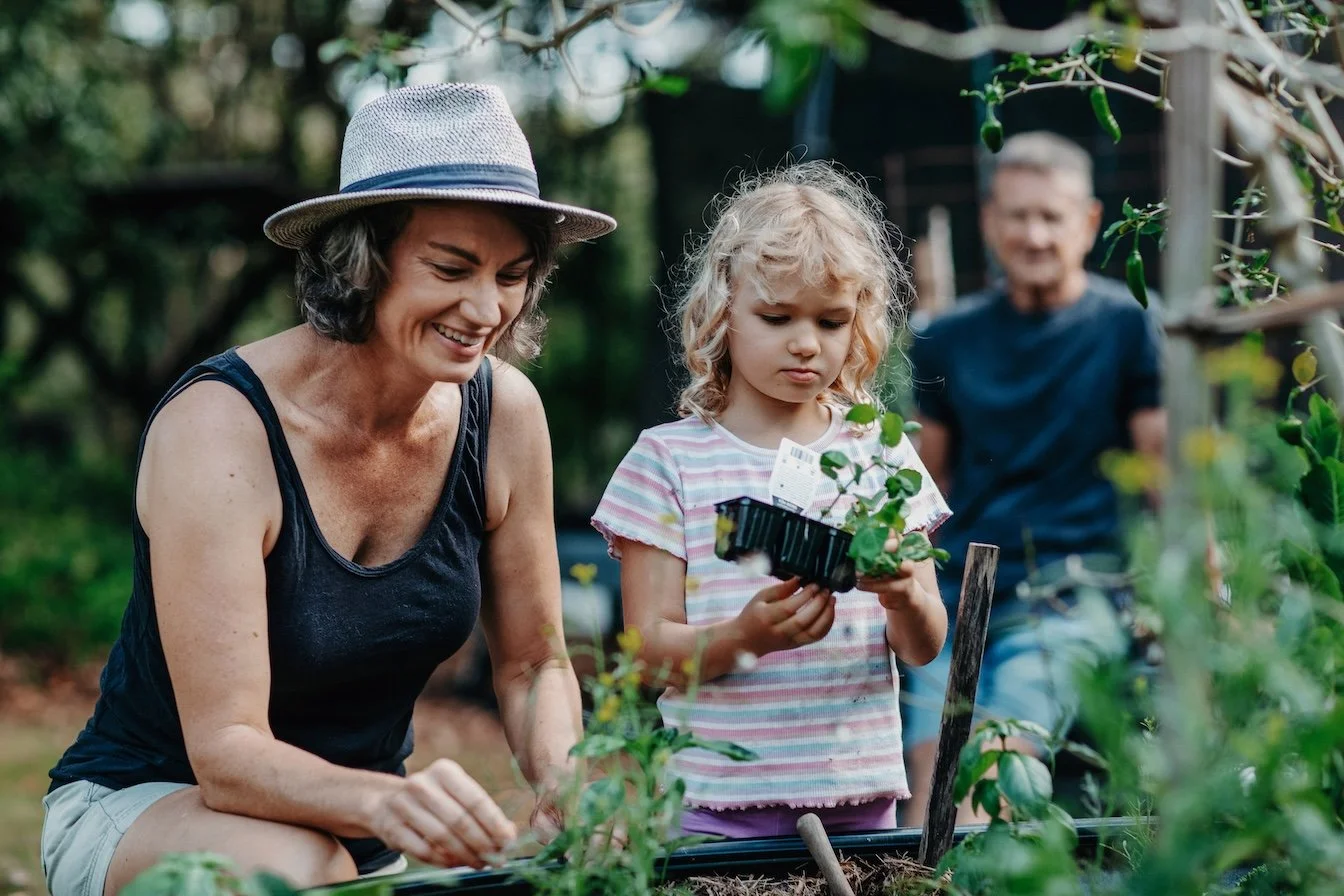 Tarla with her family outdoors. They are gardening, her daughter is holding a small plant and a tray, her husband is in the background.