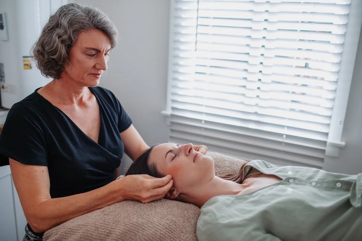 A client is lying on a massage table with her eyes closed, receiving a jaw release treatment from a Tarla, in a her treatment room in Eudlo.