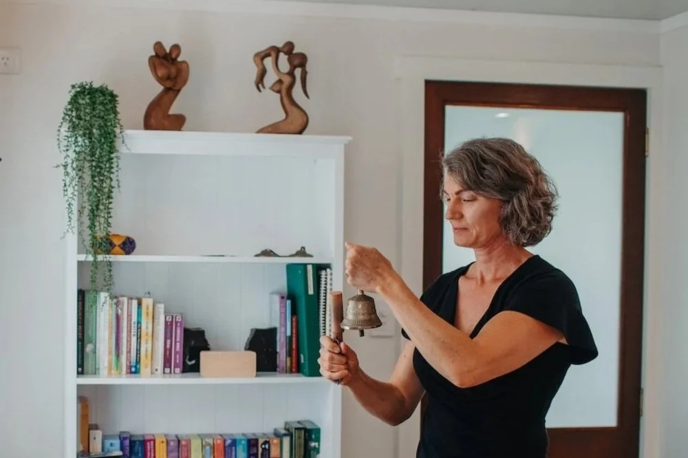 Tarla ringing a small metal bell as part of her treatment in her treatment room with a white bookshelf, decorative sculptures on top, a hanging green plant.