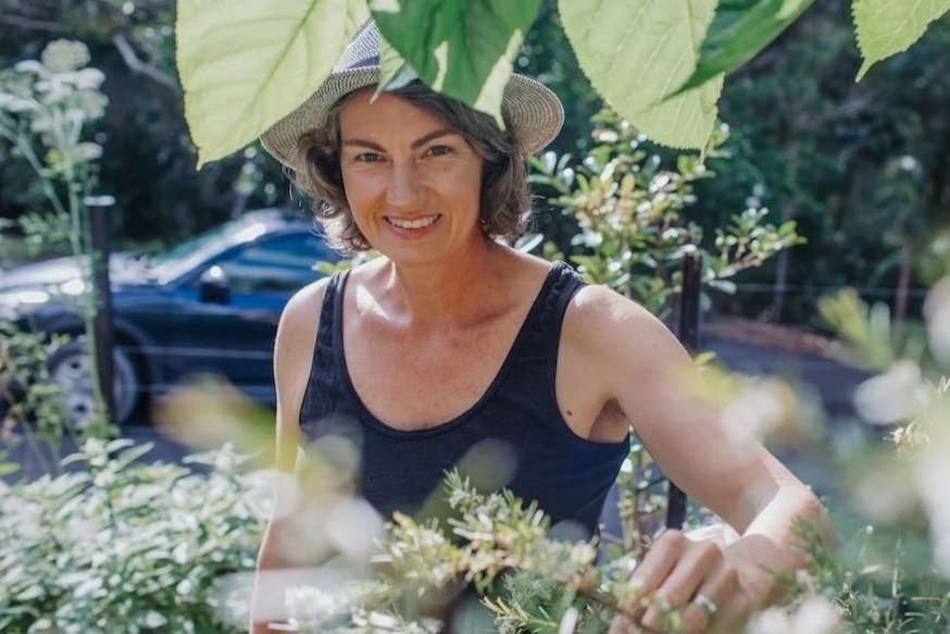 Tarla wearing a wide-brimmed hat and black tank top, smiling while gardening among green plants and leaves on a sunny day.