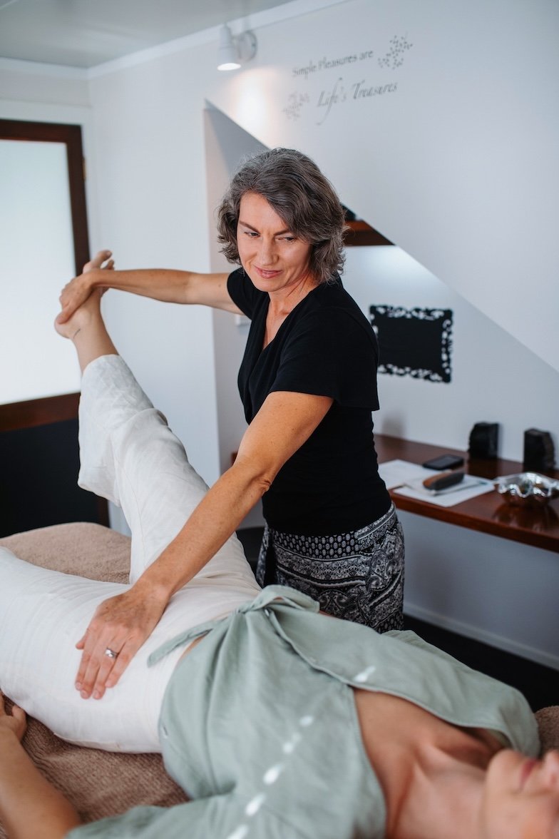 Tarla doing a Kinesiology balance on a client lying on a treatment table in her room in Eudlo with white walls and wooden furniture.