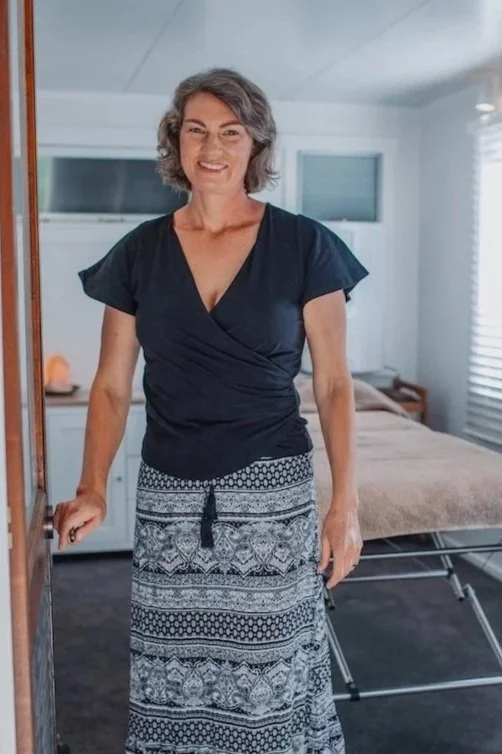 Tarla standing in the door of her treatment room, smiling at the camera. She is wearing a black V-neck top and a long patterned skirt. The room has a treatment bed, windows with blinds, and light-coloured walls.
