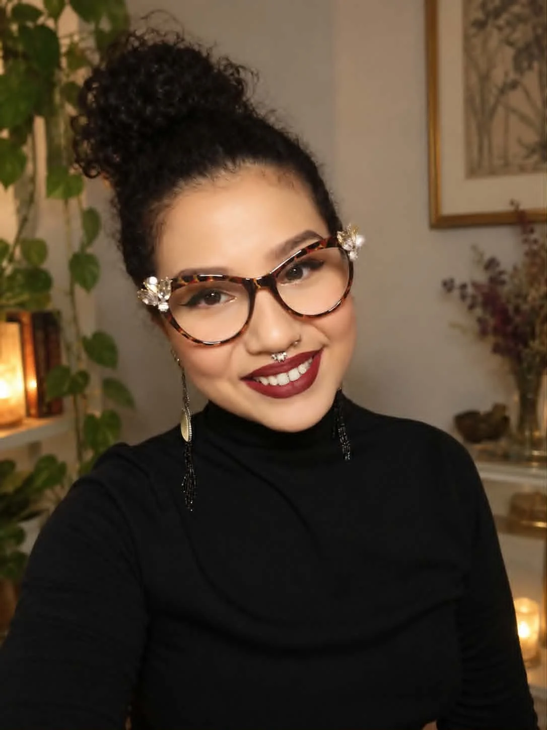 A young woman with curly hair in a bun, wearing large tortoiseshell glasses with decorative flowers, a septum nose ring, earrings, and a black turtleneck. She is smiling and standing indoors with a plant, framed artwork, and warm lighting in the background.