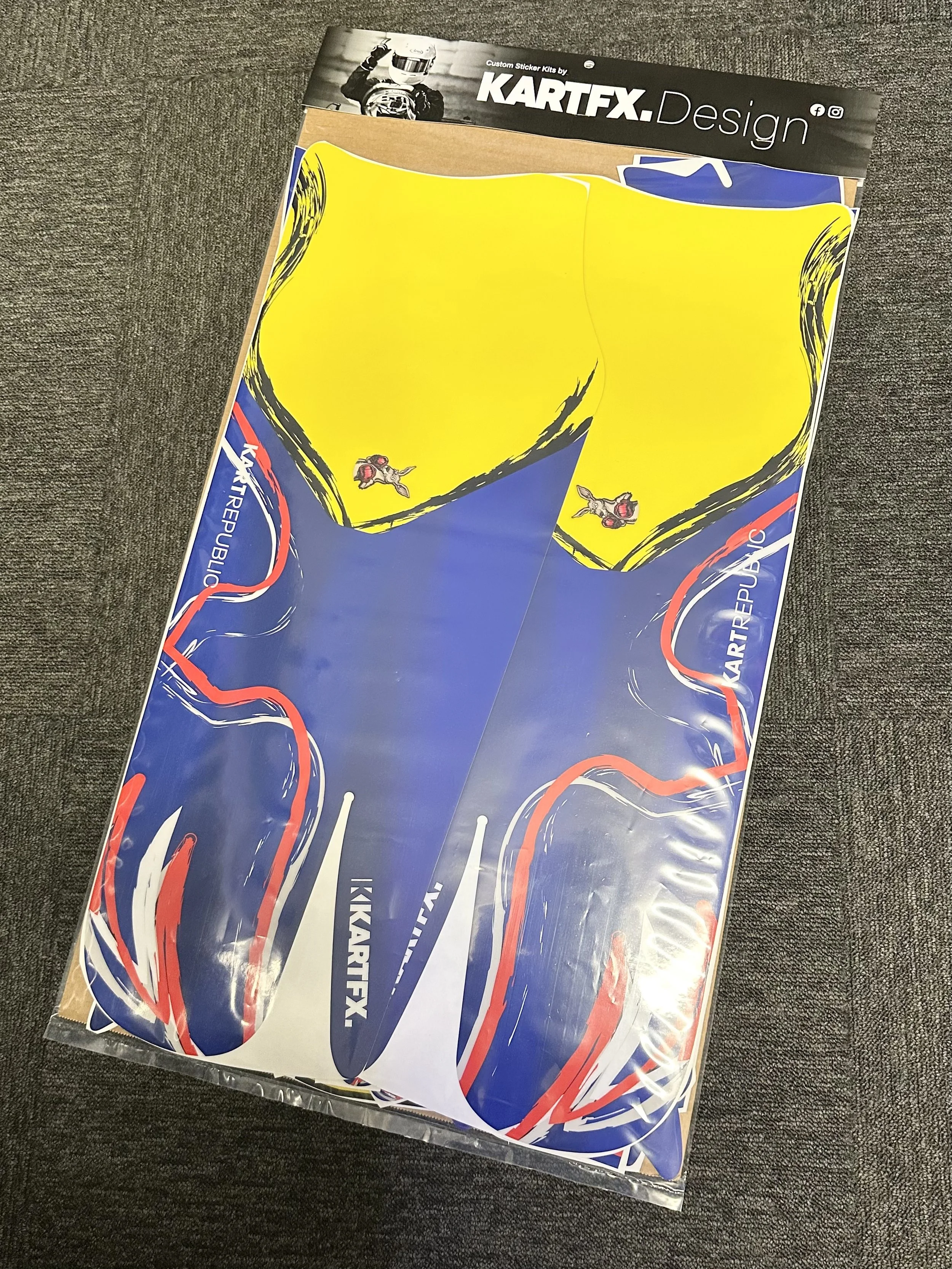 Colorful decal stickers for kart racing, featuring blue, yellow, white, and red designs, placed inside a clear plastic packaging on a dark fabric surface.