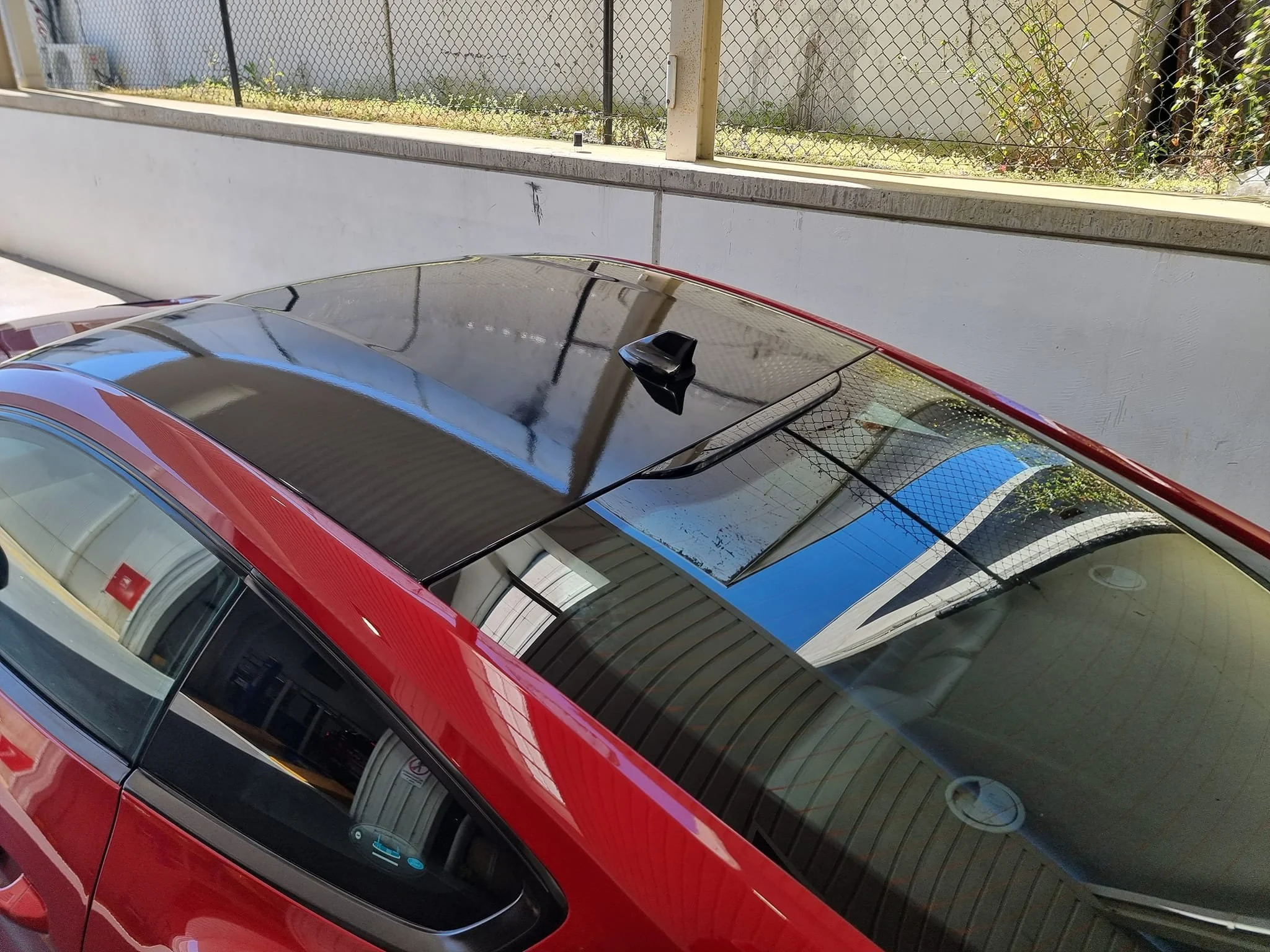Top view of a red car with a black roof parked near a white wall and a chain-link fence, showing reflections of the surroundings on the windshield and roof.
