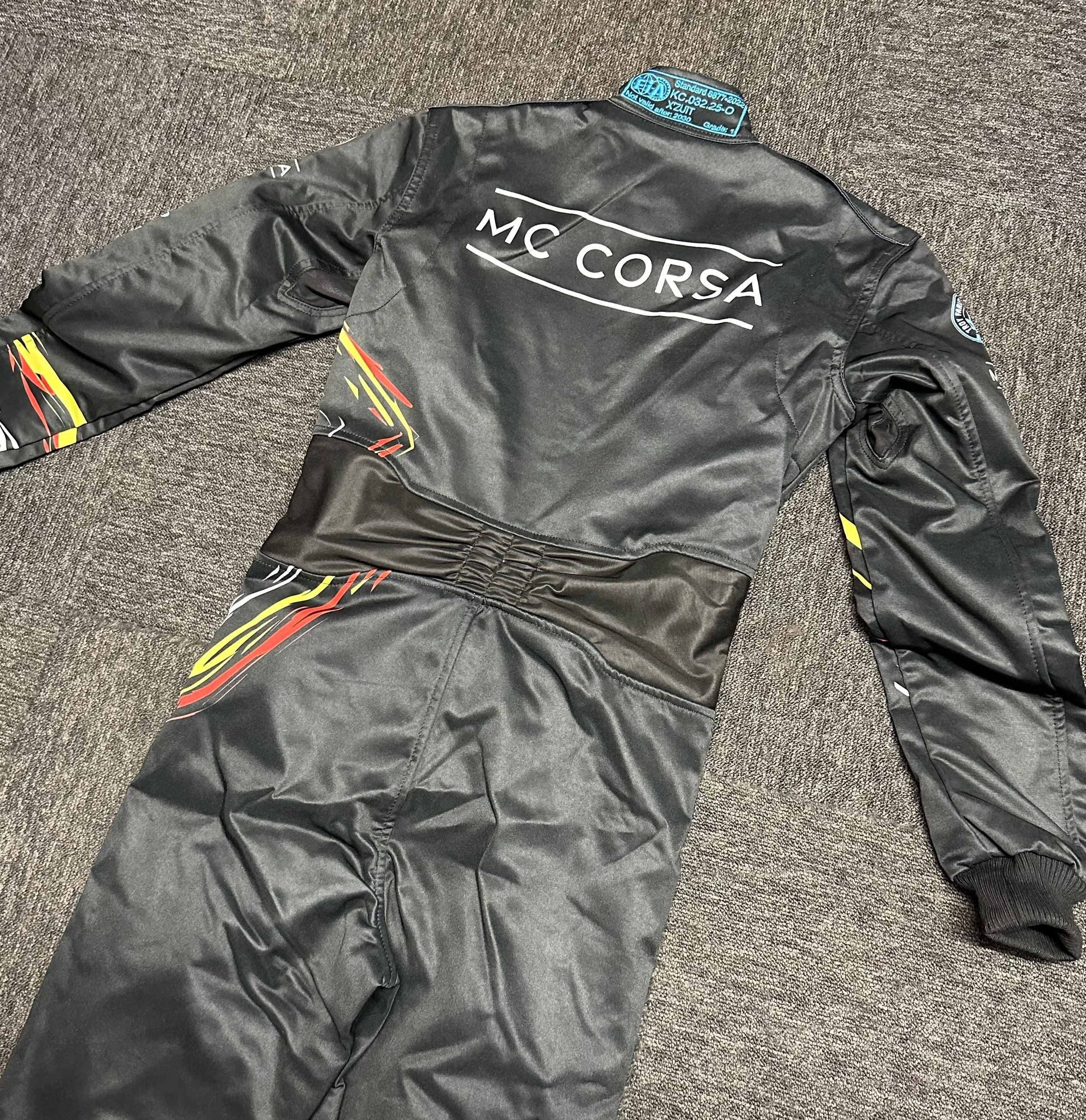 A black racing suit with colorful red, yellow, and white graphics on the arms and legs, and the words 'MC Corsa' on the back, laid out flat on a gray carpeted floor.
