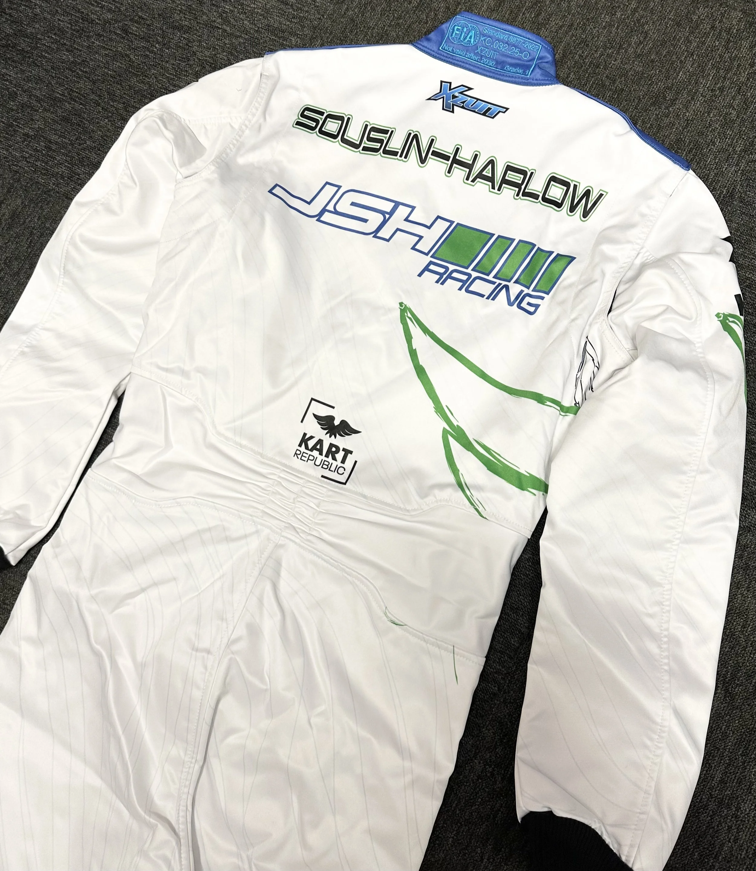 White racing suit with multiple sponsor logos and green graphics, laid flat on a dark surface.