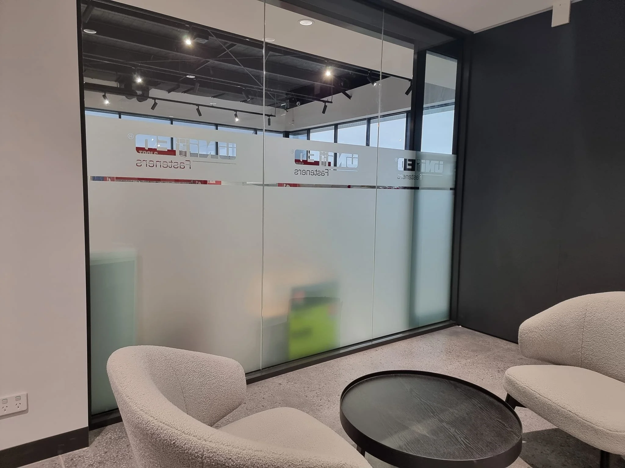 Modern office lounge with two beige chairs and a black round table in front of frosted glass walls with partial company branding, inside a contemporary office space with black ceiling and track lighting.