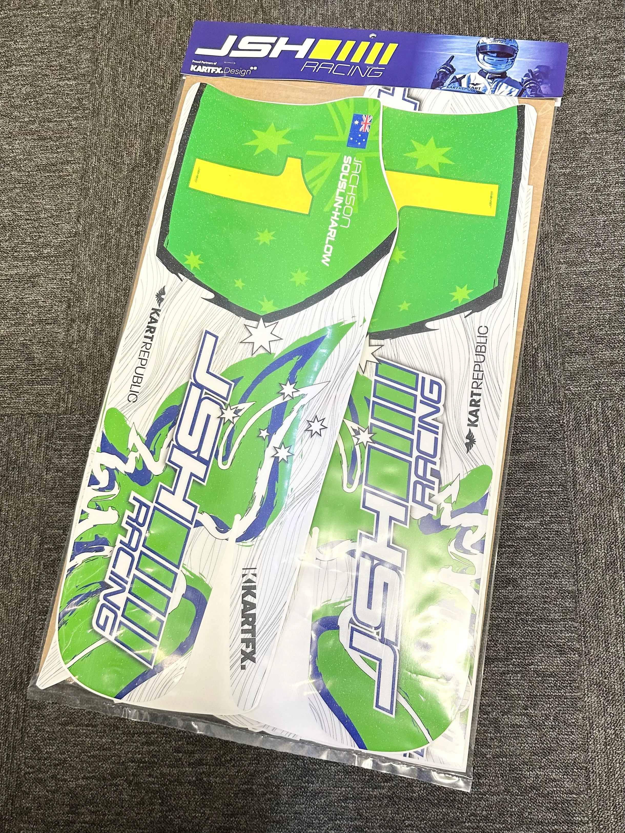 Graphics decal stickers for JSH Racing, a kart racing team, with green, white, and blue colors, featuring the team name and Australian flag, on a plastic sheet, on a gray fabric surface.