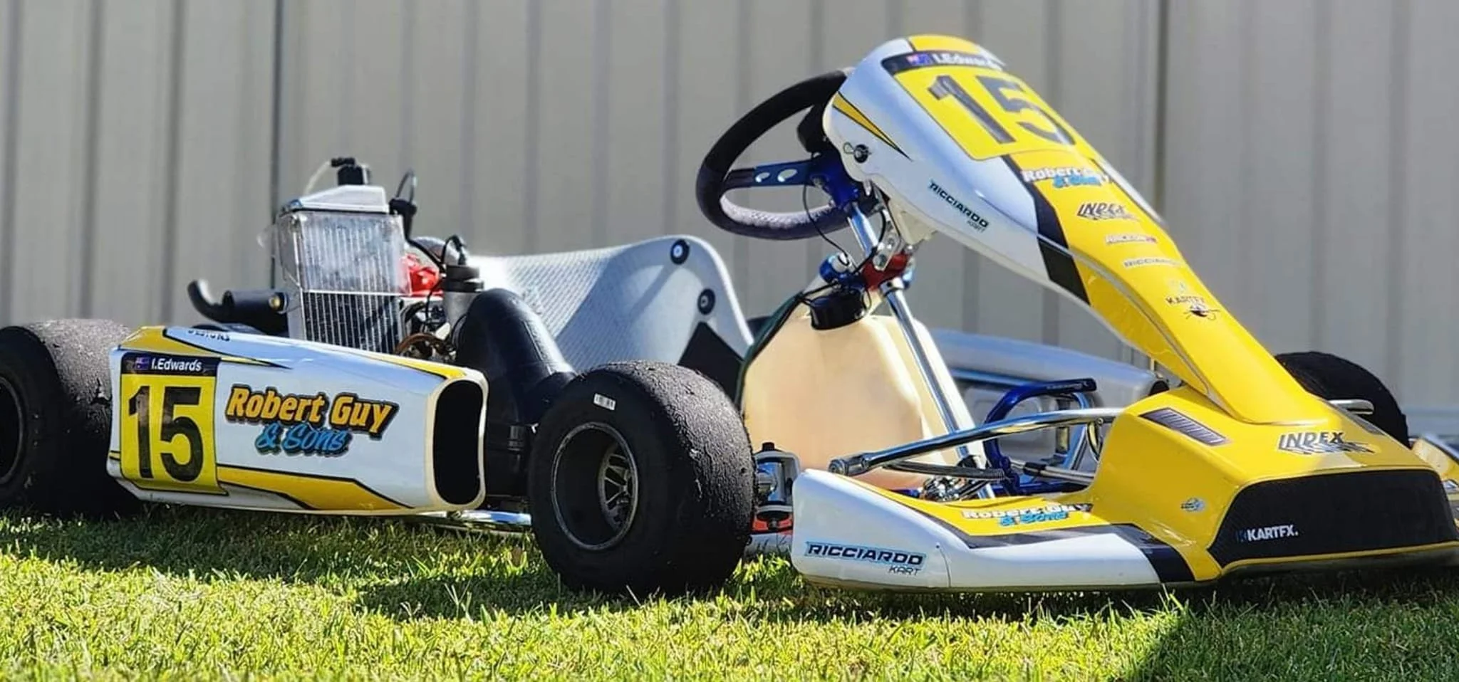 A vintage yellow and white go-kart with the number 15 on the side, labeled 'Robert Guy & Sons,' resting on a grassy area in front of a gray metallic wall.
