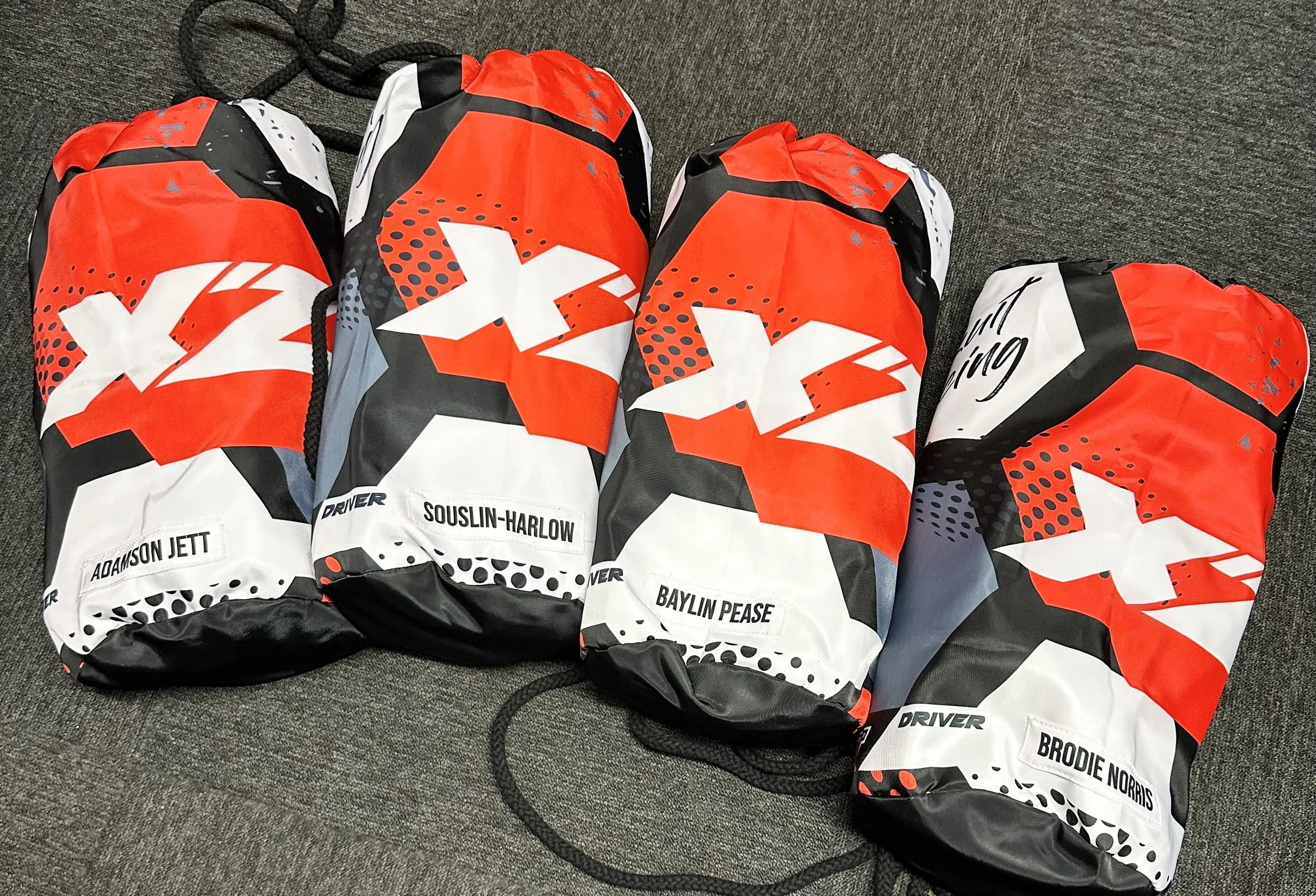 Four red, white, and black drawstring bags with a star-themed design, each labeled with a different name: Adamson Jett, Souslin-Harlow, Baylin Pease, Brodie Norris.