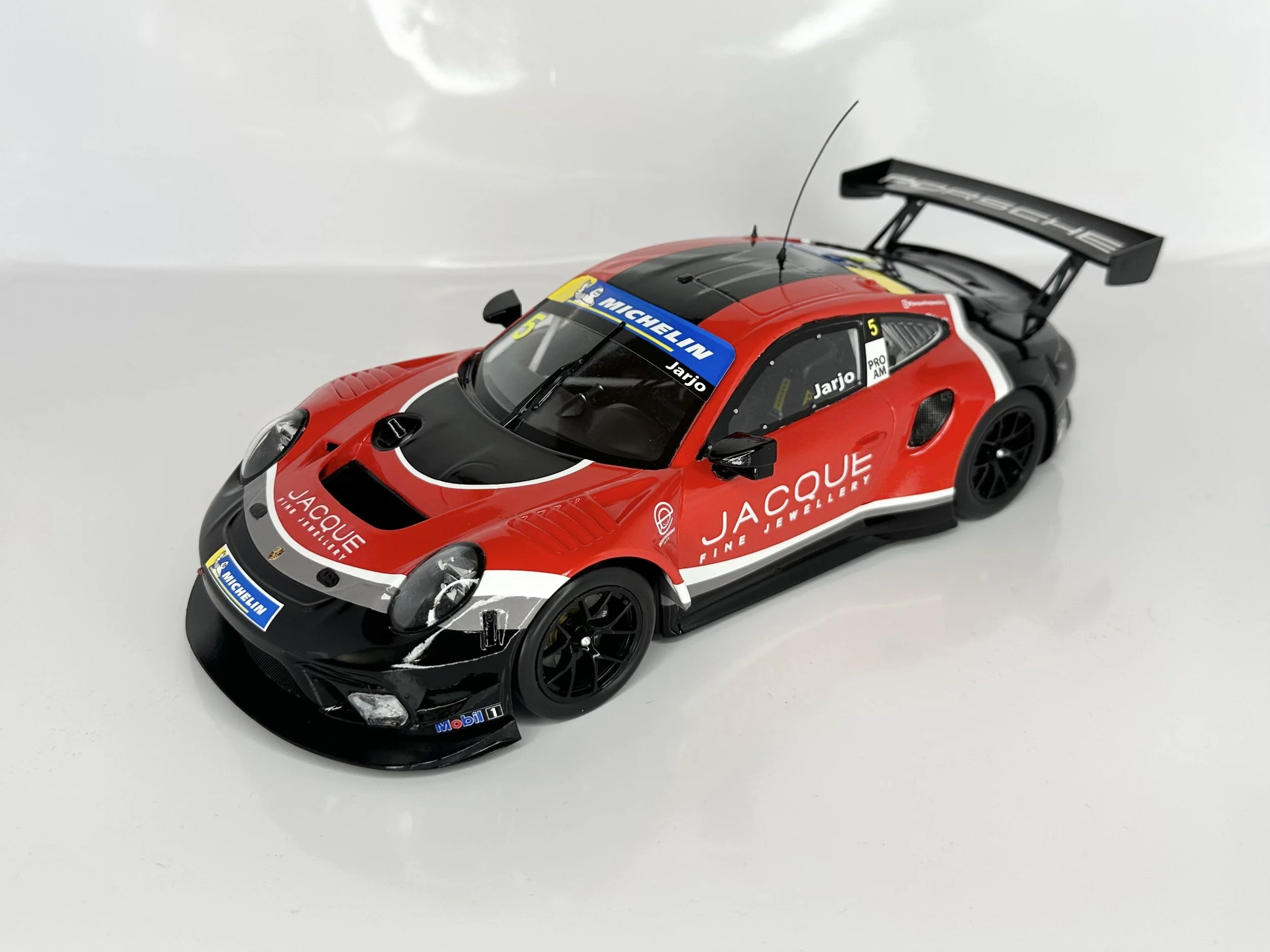 A detailed scale model of a Porsche race car with red, black, and white livery featuring sponsor logos including Jacques Fine Jewellers, Michelin, and Mobil, positioned on a light-colored surface.