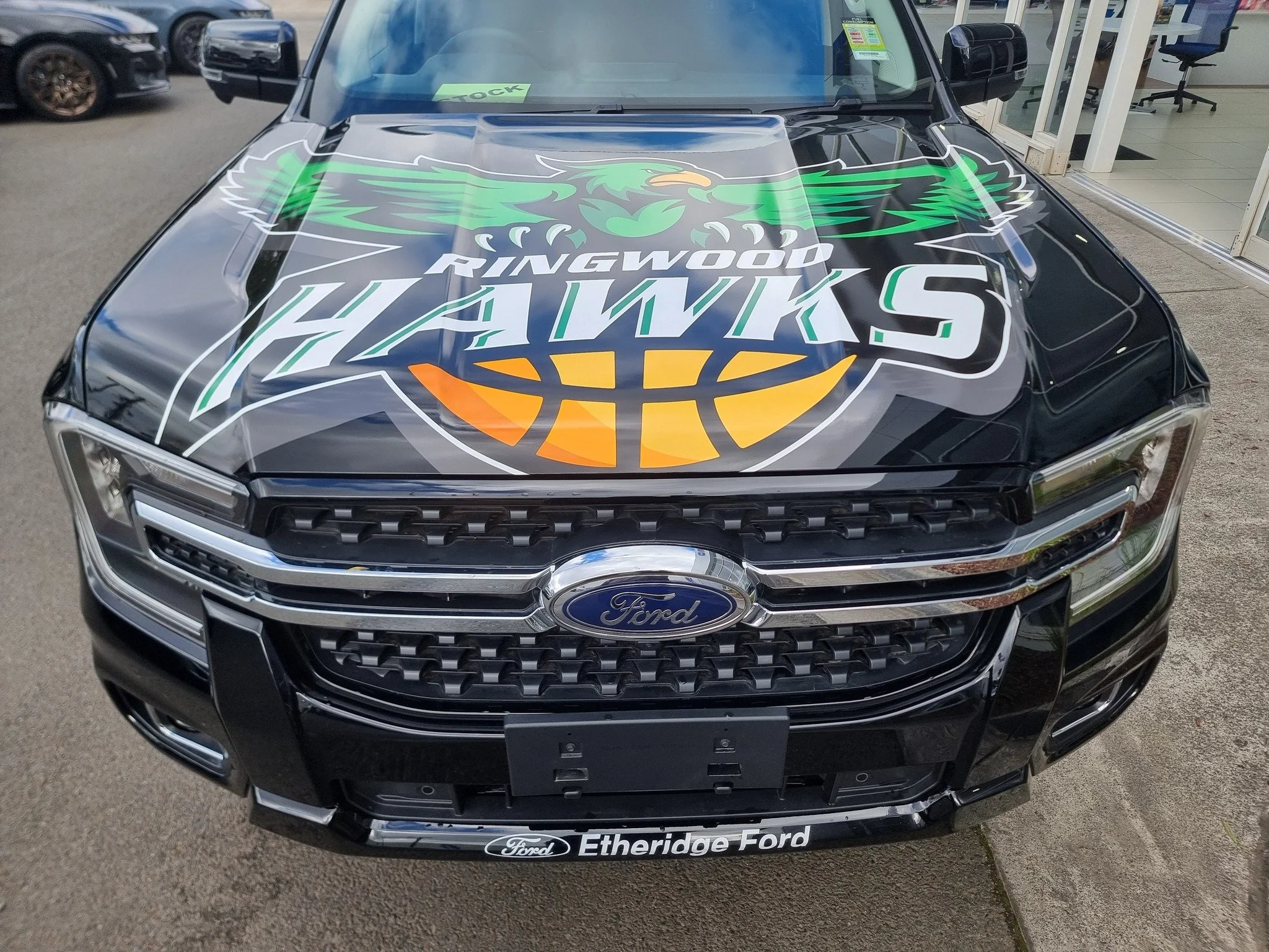 Black Ford vehicle with a large Seattle Kraken hockey team logo on the hood, featuring an eagle face with green wings and the text 'Ringwood Hawks'. The car is parked outside a dealership with a sign that reads 'Etheridge Ford'.