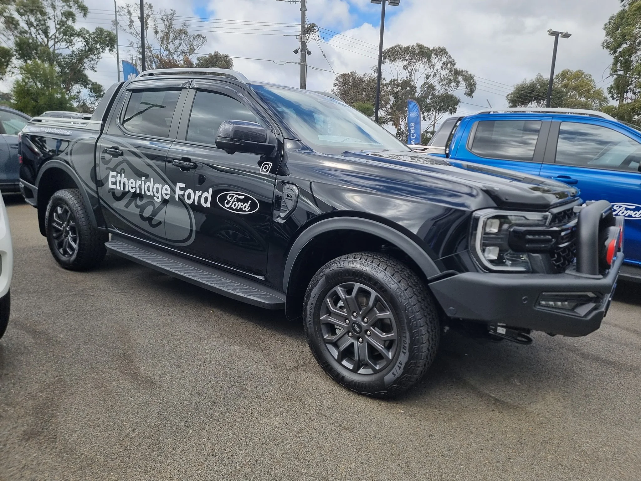 Black Ford pickup truck with 'Etheridge Ford' branding and logo, parked at a dealership lot with other blue and white vehicles visible in the background.