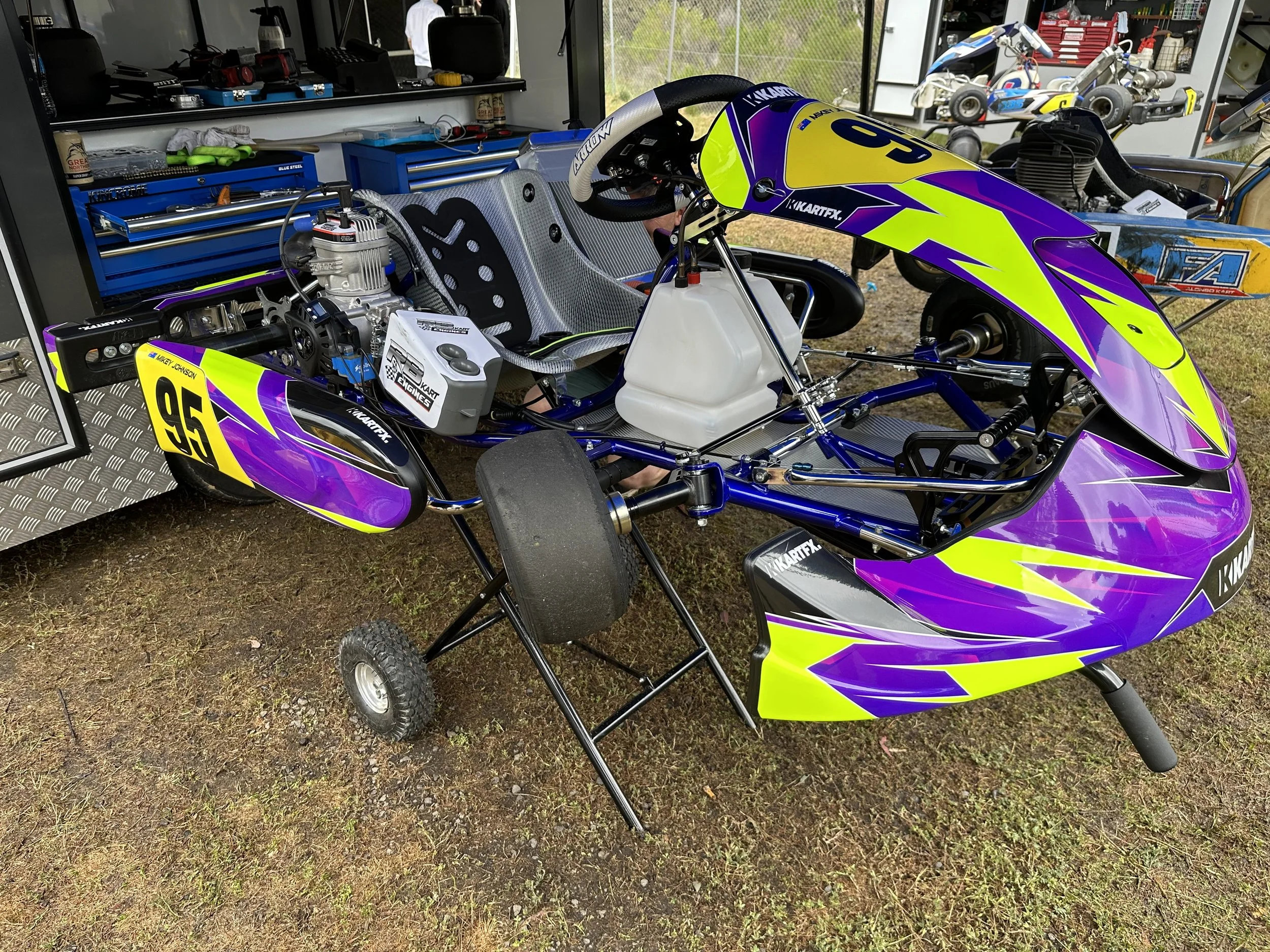 A go-kart with a purple, yellow, and black body, number 95, on a grassy area, with tools and equipment in a nearby garage in the background.