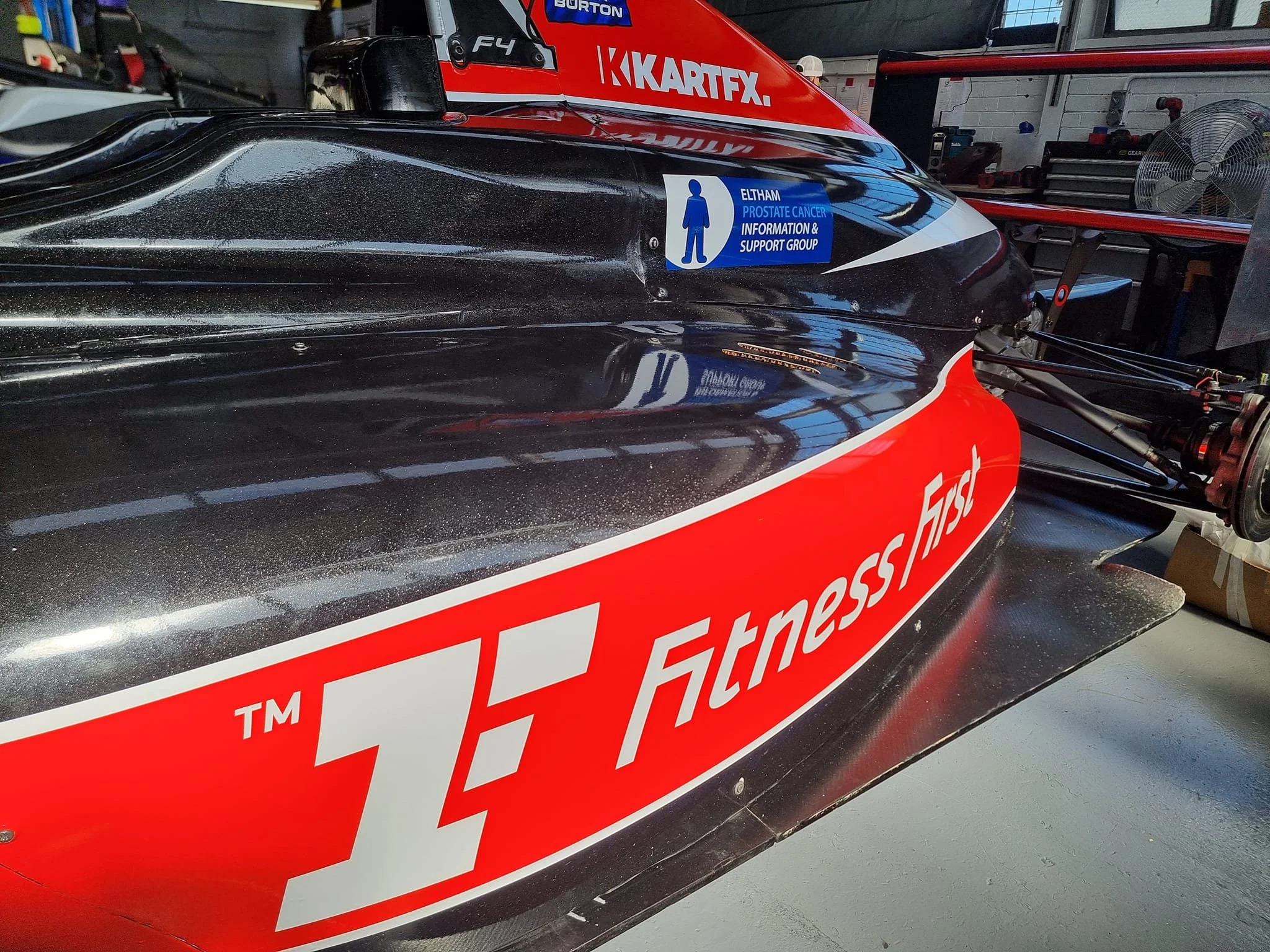 Close-up of a race car's nose with a black and red paint job, featuring sponsor logos including 'Atness.first' and a sticker supporting prostate cancer awareness.
