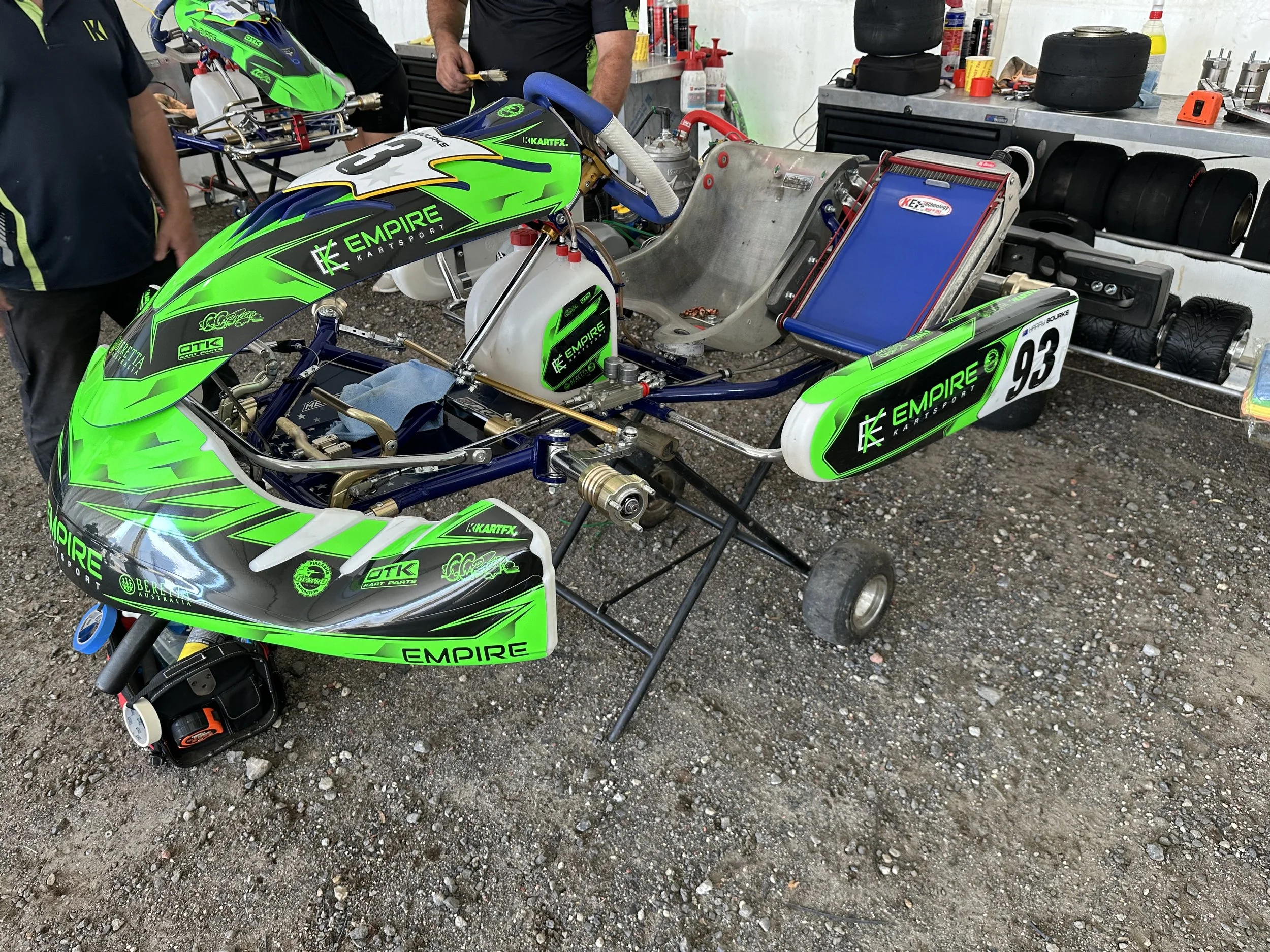 A go-kart with green, black, and white graphics labeled 'Empire Karting' and the number 93, placed on a gravel surface in a racing paddock. The go-kart is in a maintenance area with tires, tools, and equipment in the background.
