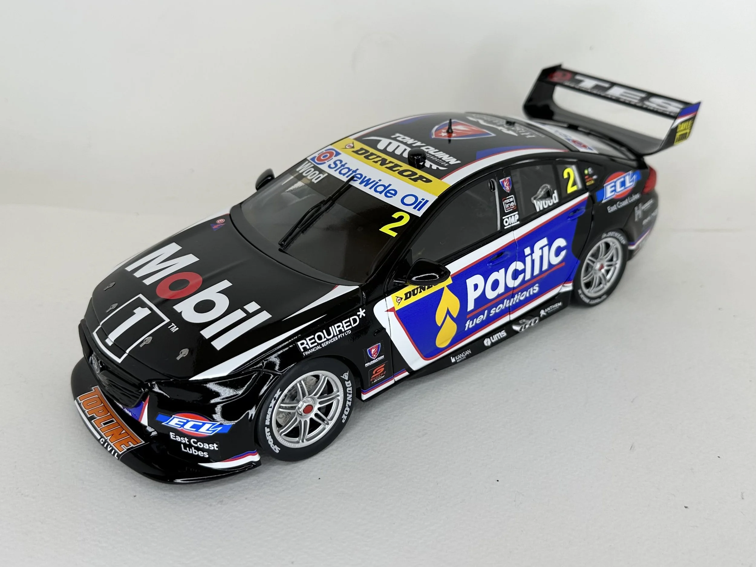 A detailed scale model of a racing car with black, blue, and white livery, featuring sponsor logos such as Mobil 1, Pacific Fuel Solutions, and East Coast Lubes, positioned on a plain white surface.