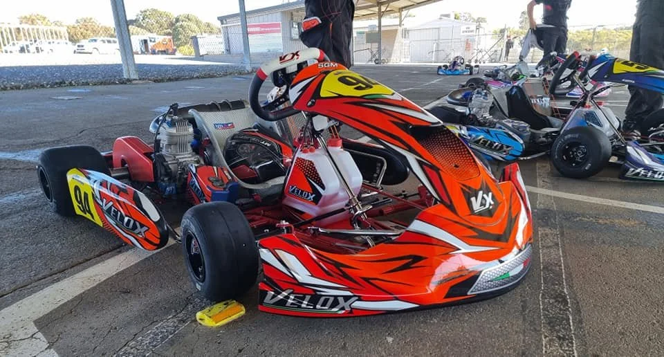 Red and orange go-kart with black tires, parked under a shelter with other go-karts and people in the background.