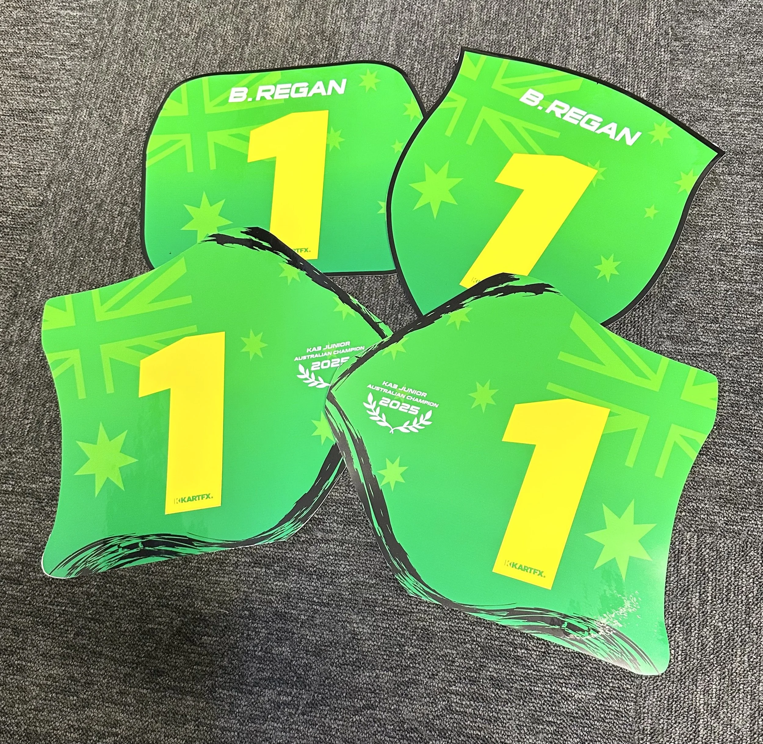 Four green and yellow kart racing trophies with the number 1 and the name B. REGAN, placed on a gray textured surface.