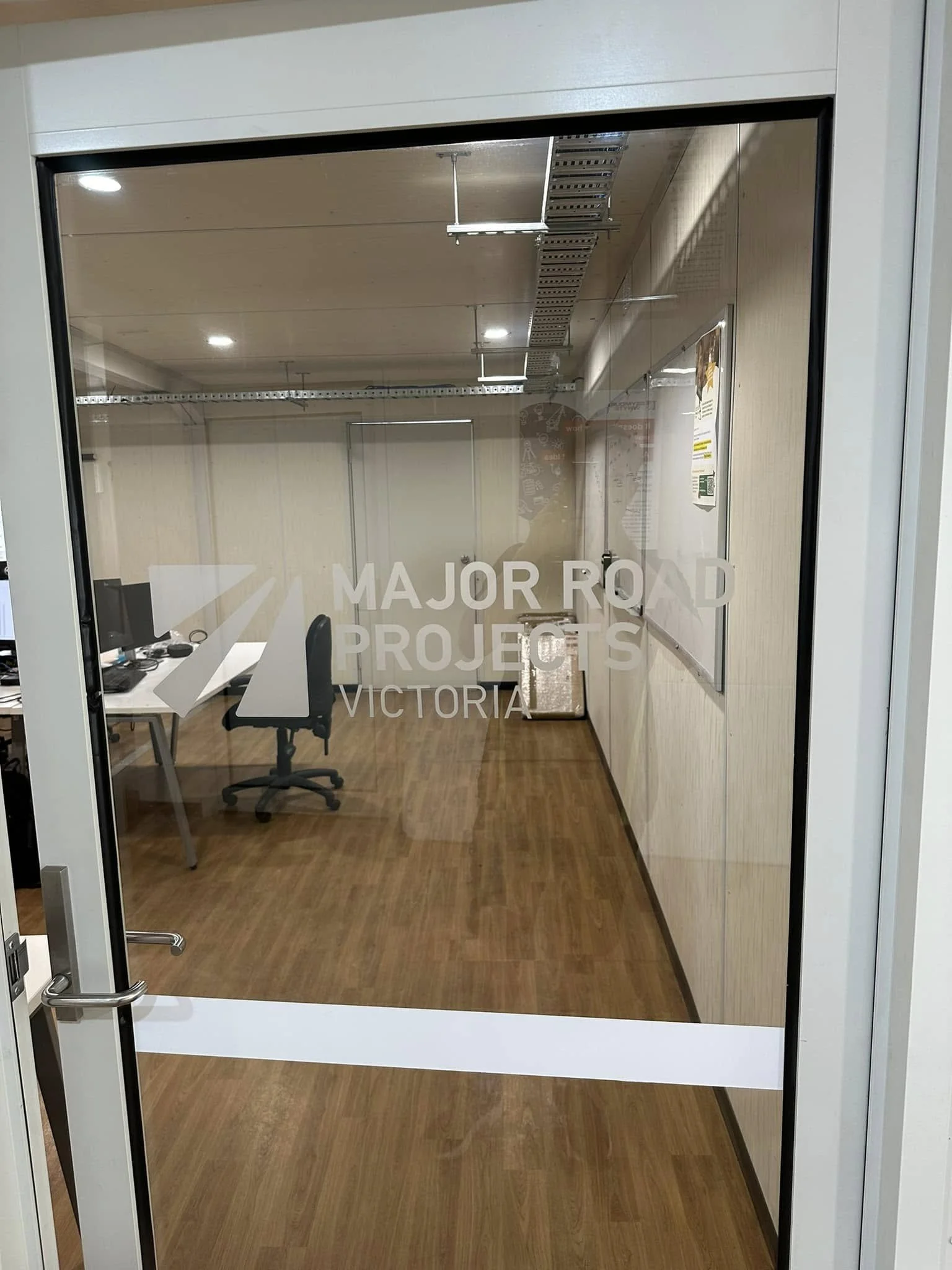 Inside an office room viewed through a glass door with 'Major Road Projects Victoria' written on it, showing desks, chairs, a whiteboard, and part of the ceiling with lighting.
