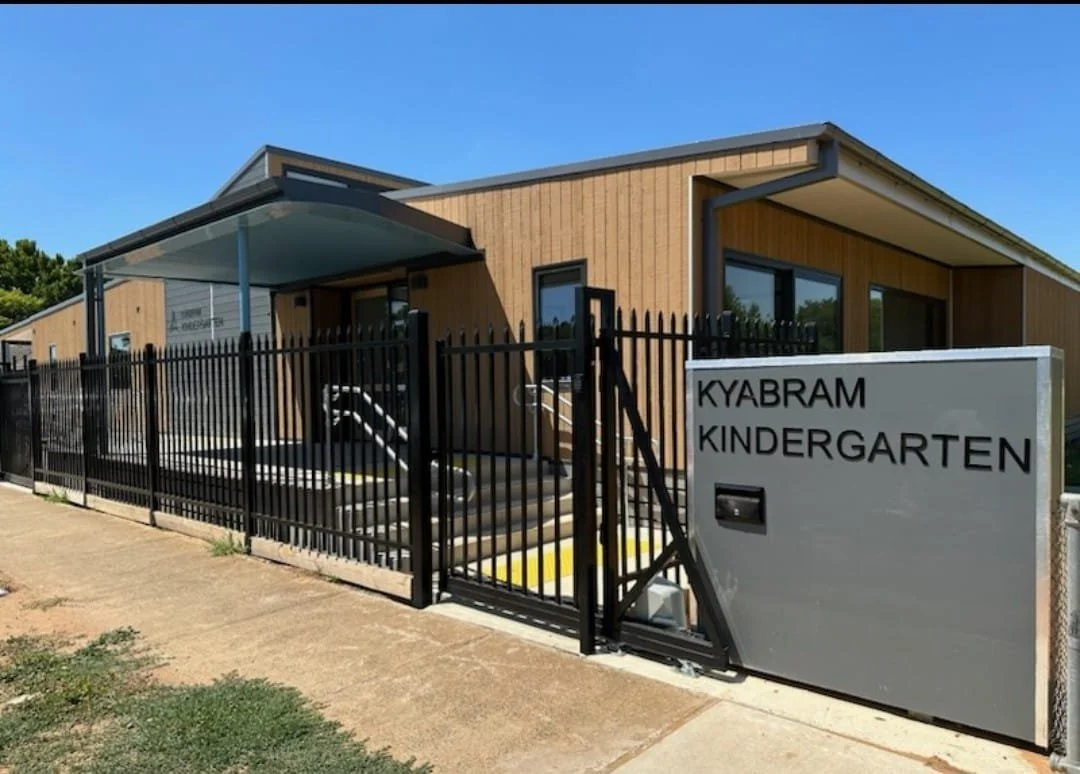 Exterior view of Kyabram Kindergarten building with a black fence, sign, and an outdoor staircase, under a clear blue sky.
