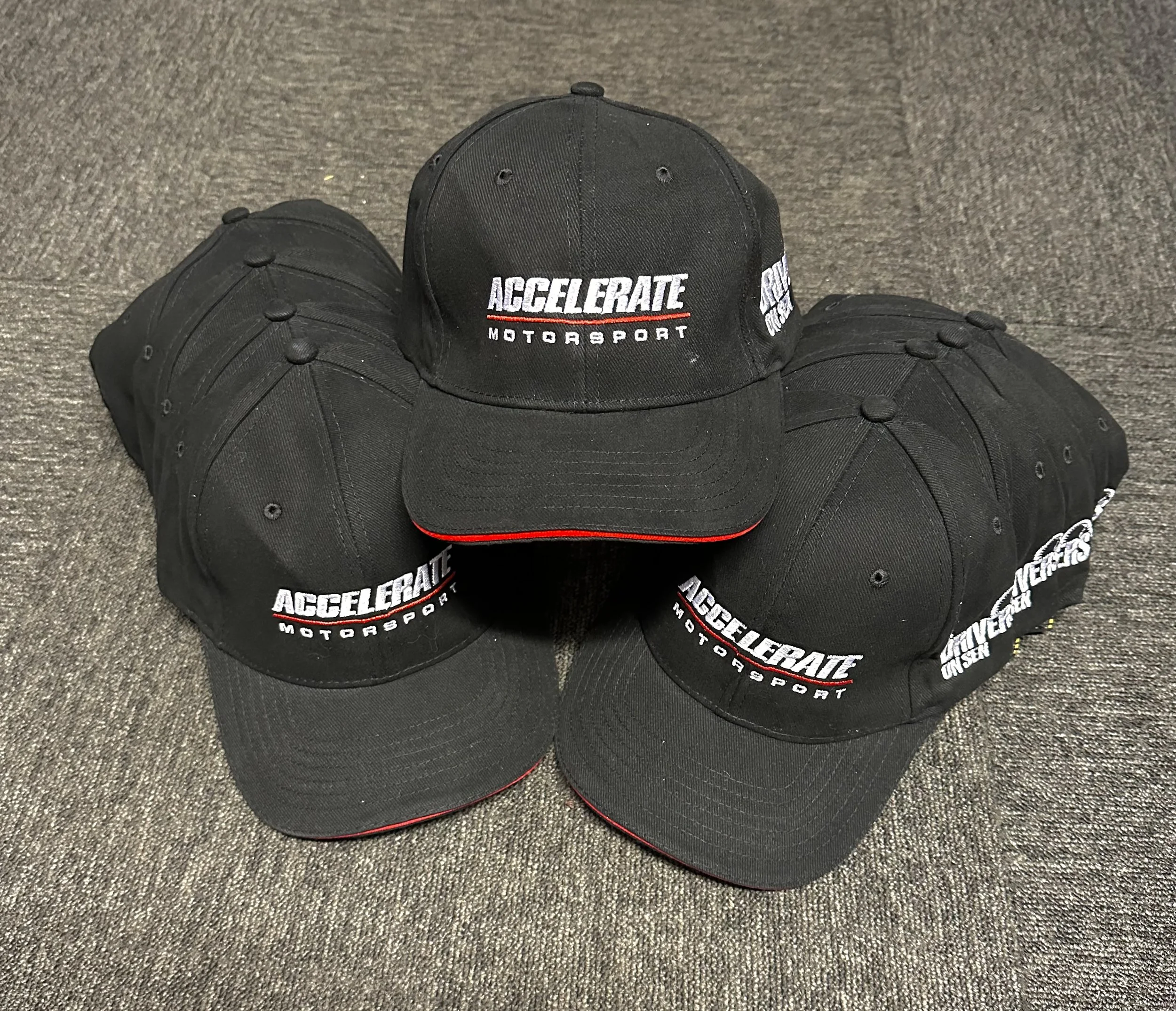 A collection of black baseball caps with white and red embroidered text that says 'Accelerate Motorsport,' arranged on a gray carpeted floor.
