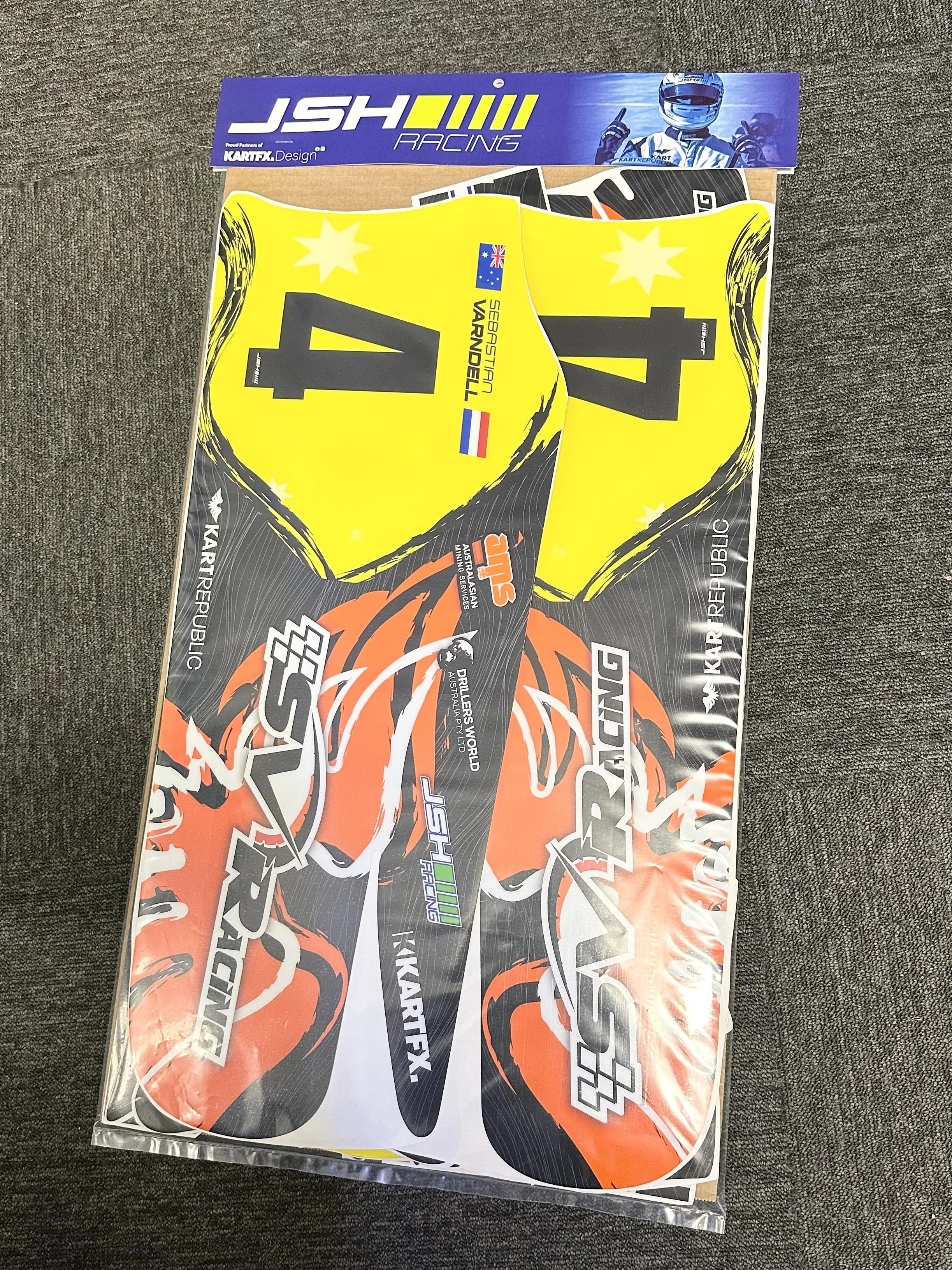 Colorful decal sheet with black, yellow, orange, and white graphics and logos, including the words 'KART RACING' and 'JSH Racing', designed for a go-kart, placed on a gray carpet.