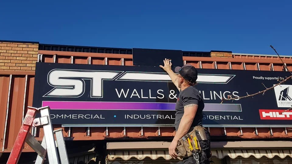 A man installing or repairing a sign for ST Walls & Coatings on a building exterior. The sign includes the company name and categories like commercial, industrial, and residential. The man is wearing a black shirt, a cap, and has tools in his belt. A