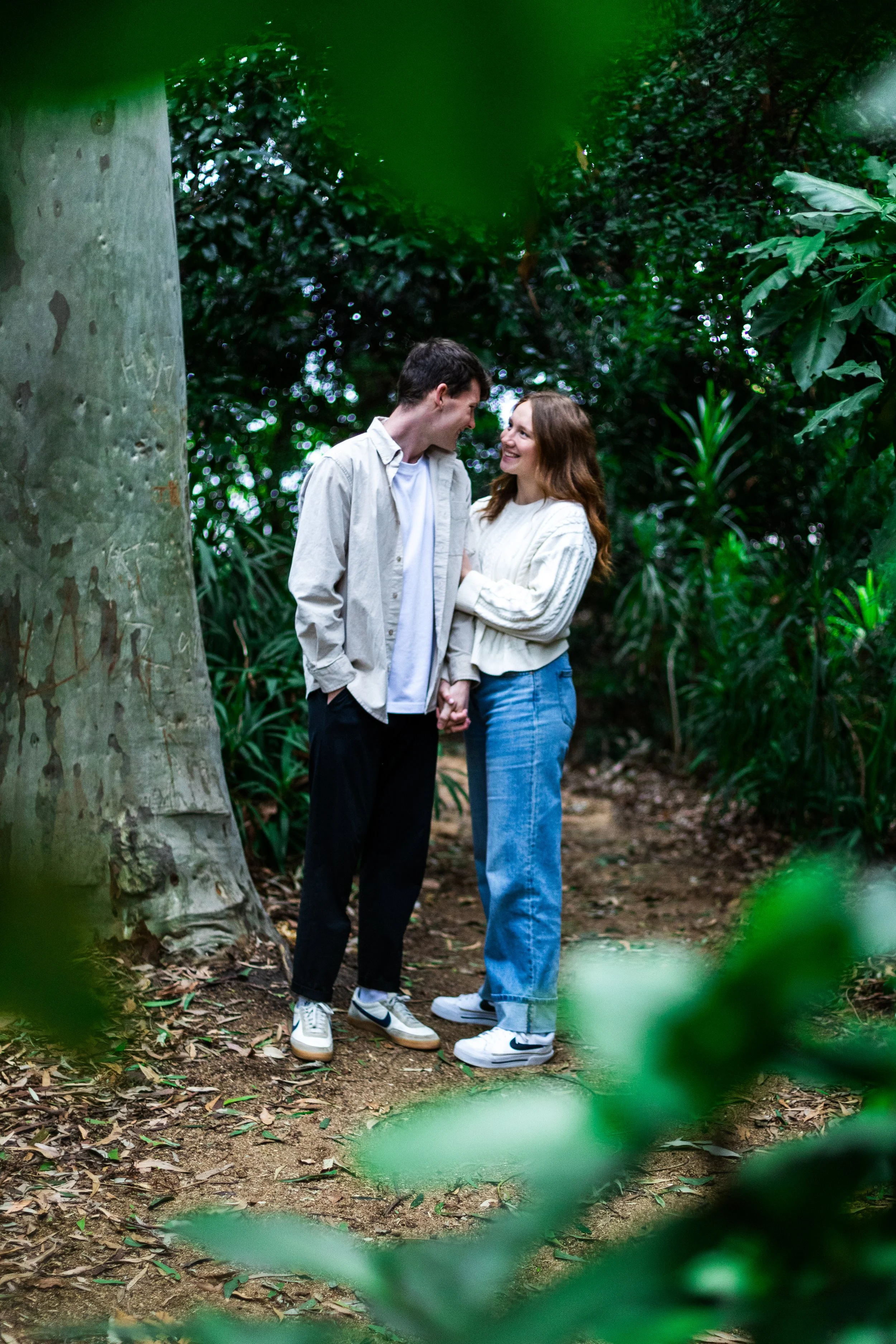 A young couple standing close together, holding hands, in a lush green forest, smiling and looking at each other affectionately.