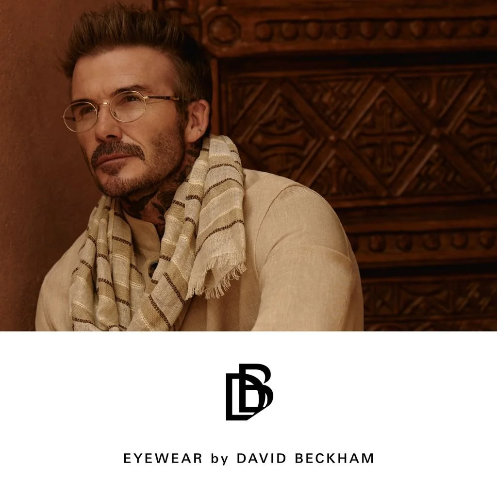 A man wearing beige clothing, glasses, and a striped scarf, sitting in front of a wooden background, with a logo for EYEWEAR by DAVID BECKHAM and branding underneath.