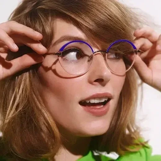 A woman with light skin, reddish-brown hair, and glasses with blue light covers, holding her glasses, available at Torquay Eye Care, with both hands, looking to her right.