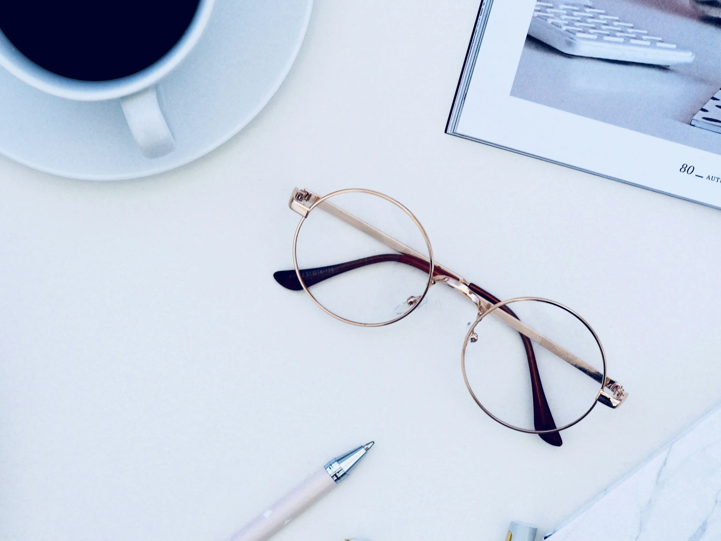 A white desk with a cup of black coffee on a saucer, a pair of round gold eyeglasses with dark brown temples, a white pen with a silver tip, and an open magazine or book with a visible page number 80 and an image of a beige keyboard.