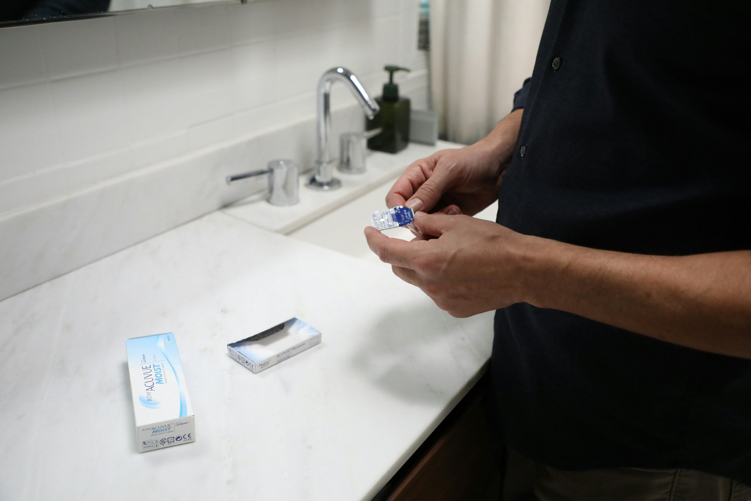 Person holding a contact lens package in a bathroom with a white countertop and a sink, and an open box of contact lenses on the counter.