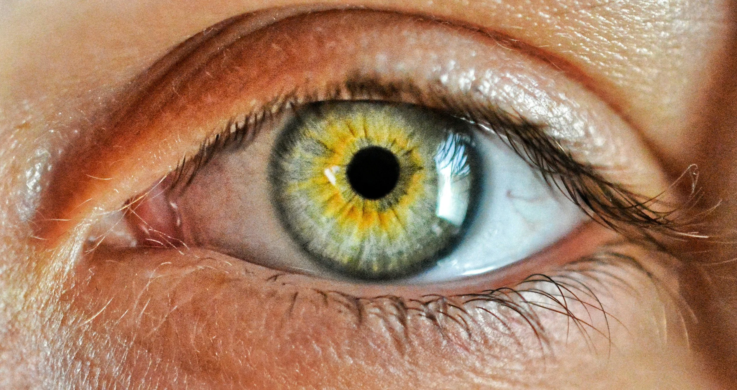 Close-up of a human eye with a green and yellow iris, detailed eyelashes, and surrounding skin.