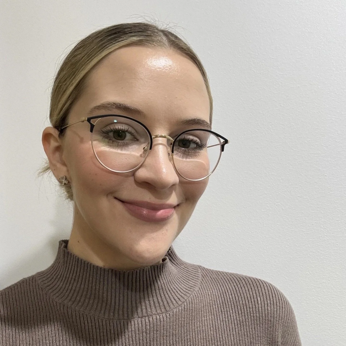 A young woman with blonde hair, wearing rounded glasses and a brown ribbed turtleneck sweater, smiling and standing against a plain white wall.